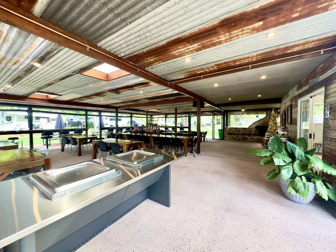 Undercover entertainment area known as the Camp Kitchen at Esk Caravan Park, featuring accessible BBQ's, pizza ovens, rustic tables, a small stage with musicians performing, warm lighting and rustic décor with wooden and brick accents. Social space.