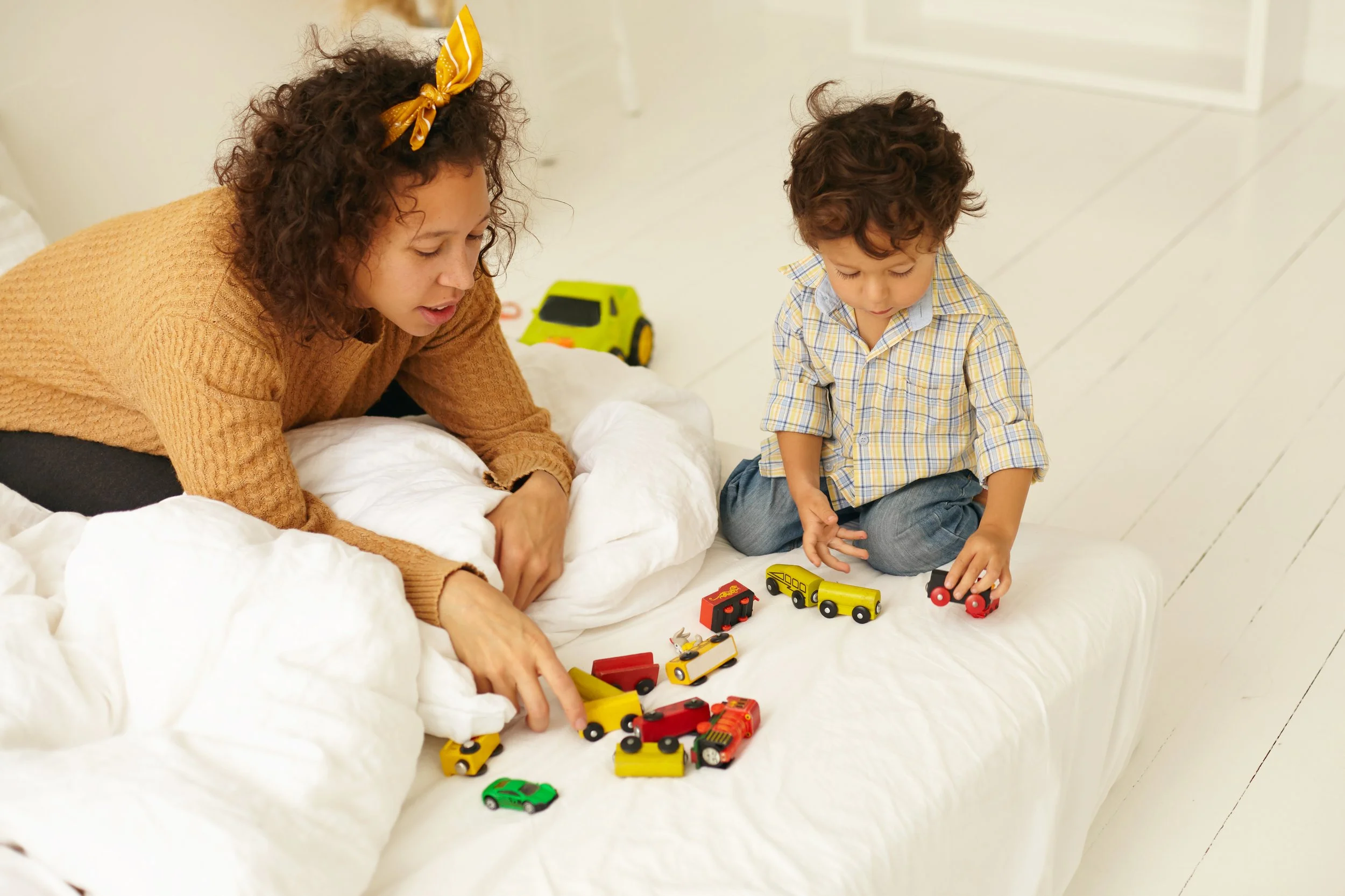 early-development-childhood-maternity-indoor-shot-curious-cute-infant-child-sitting-floor-playing-with-many-colorful-toy-cars-white-bed-sheet-mother-him-watching-him-play.jpg