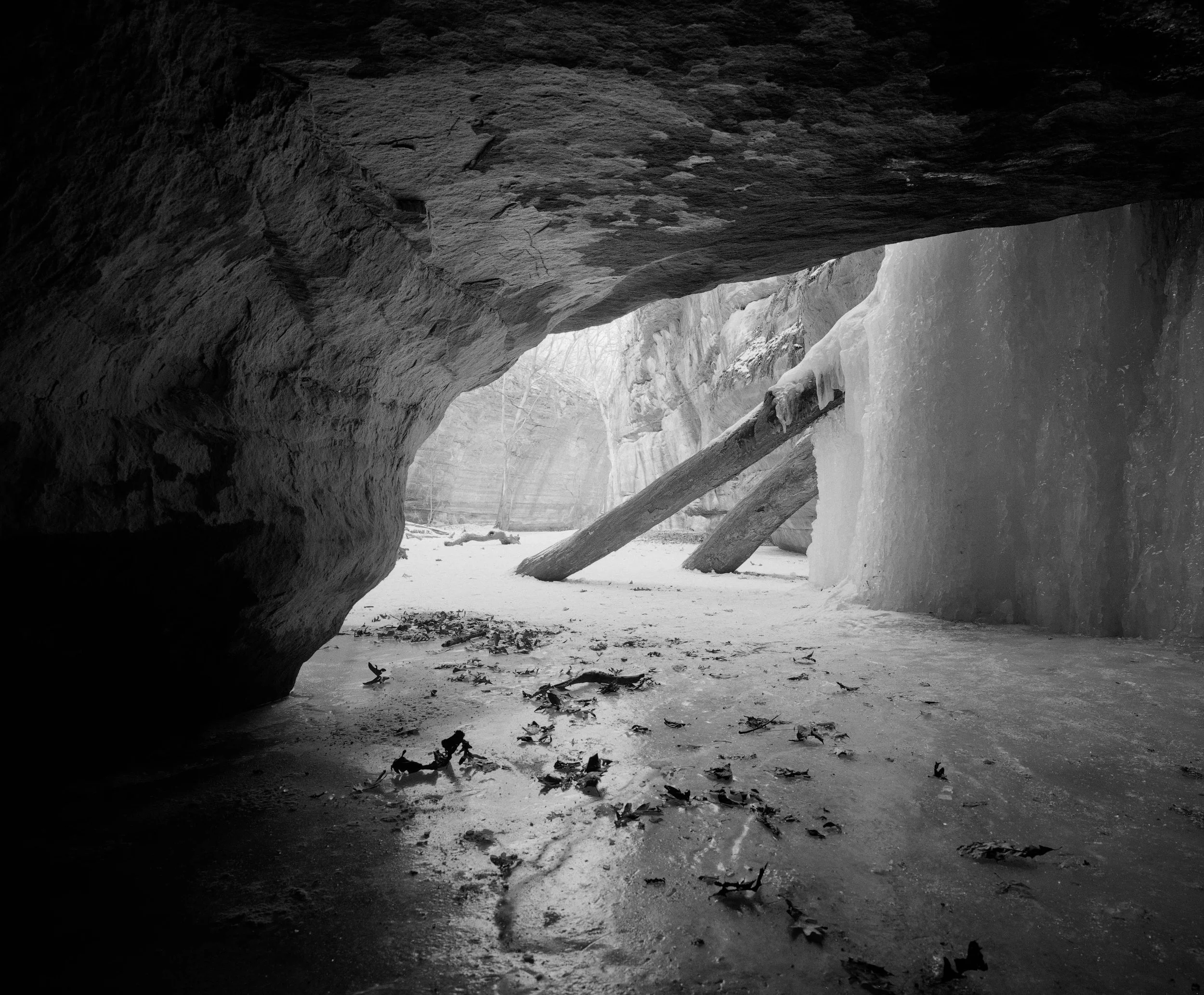 A black-and-white photo of a snowy ice cave with ice formations and fallen tree trunks on the ground.