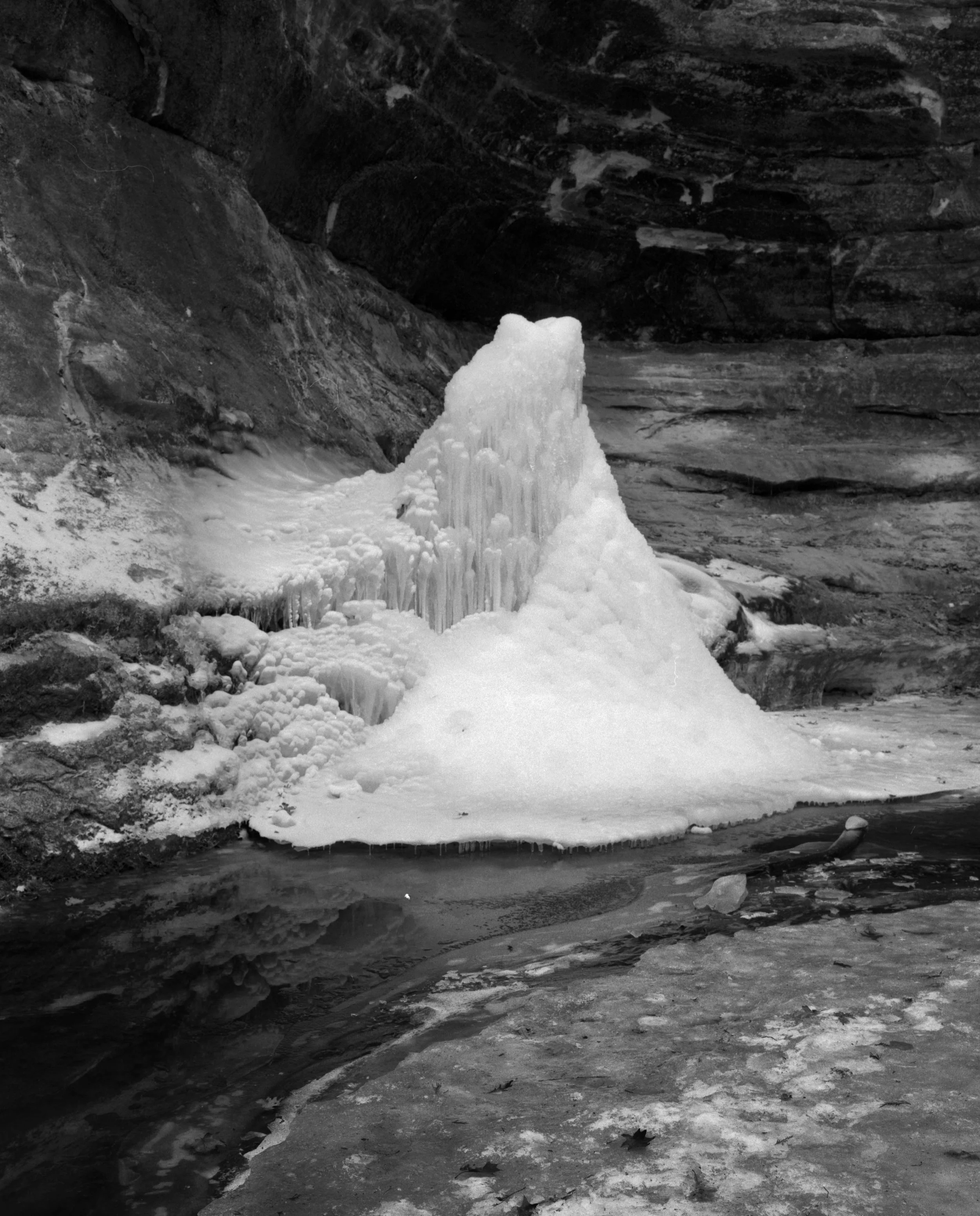 Frozen water formation in a rocky cave or crevice, with icicles and ice buildup on the surface, black and white photograph.