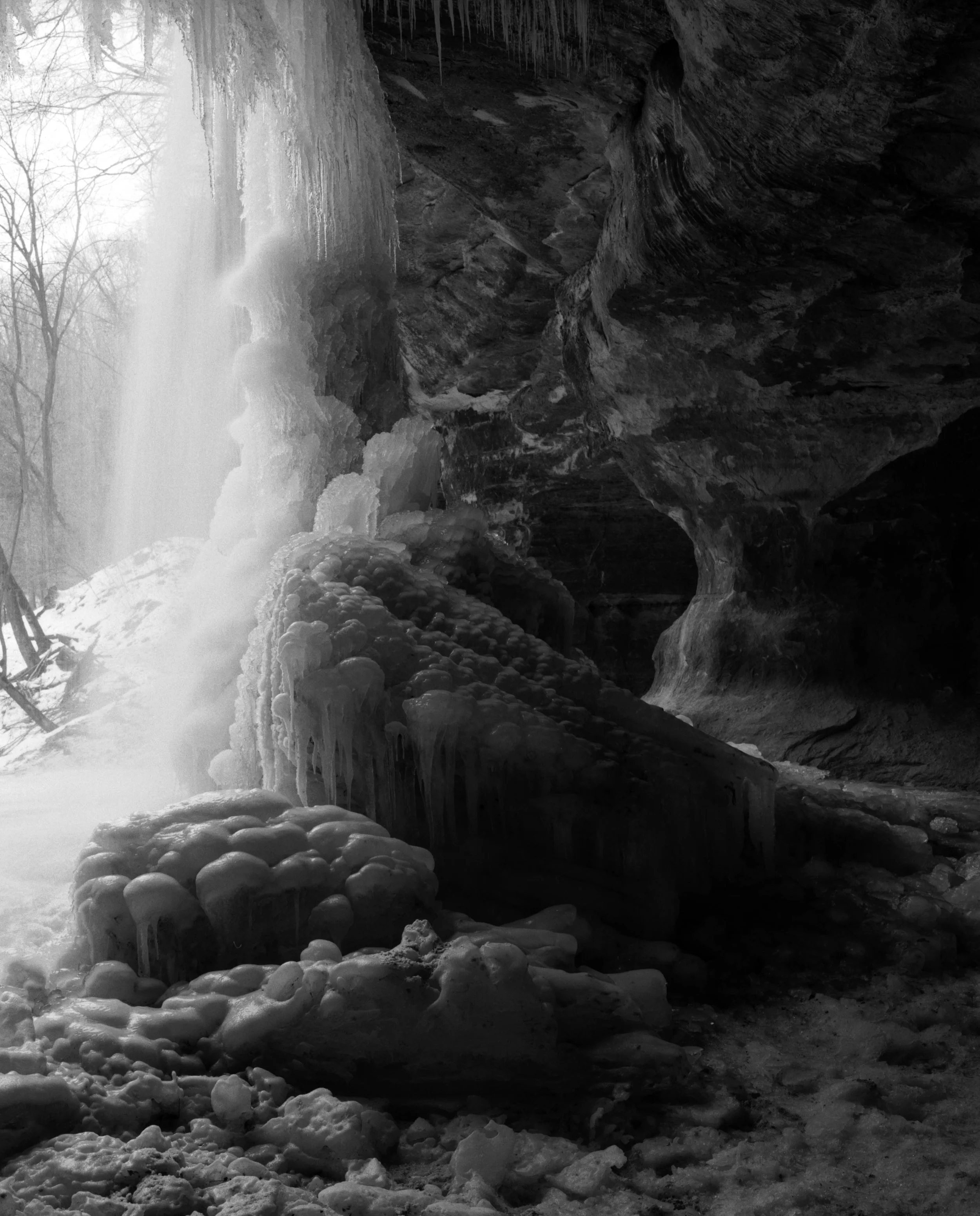 Frozen waterfall with icicles and ice formations in a rocky cave, snow-covered trees visible outside in the background.