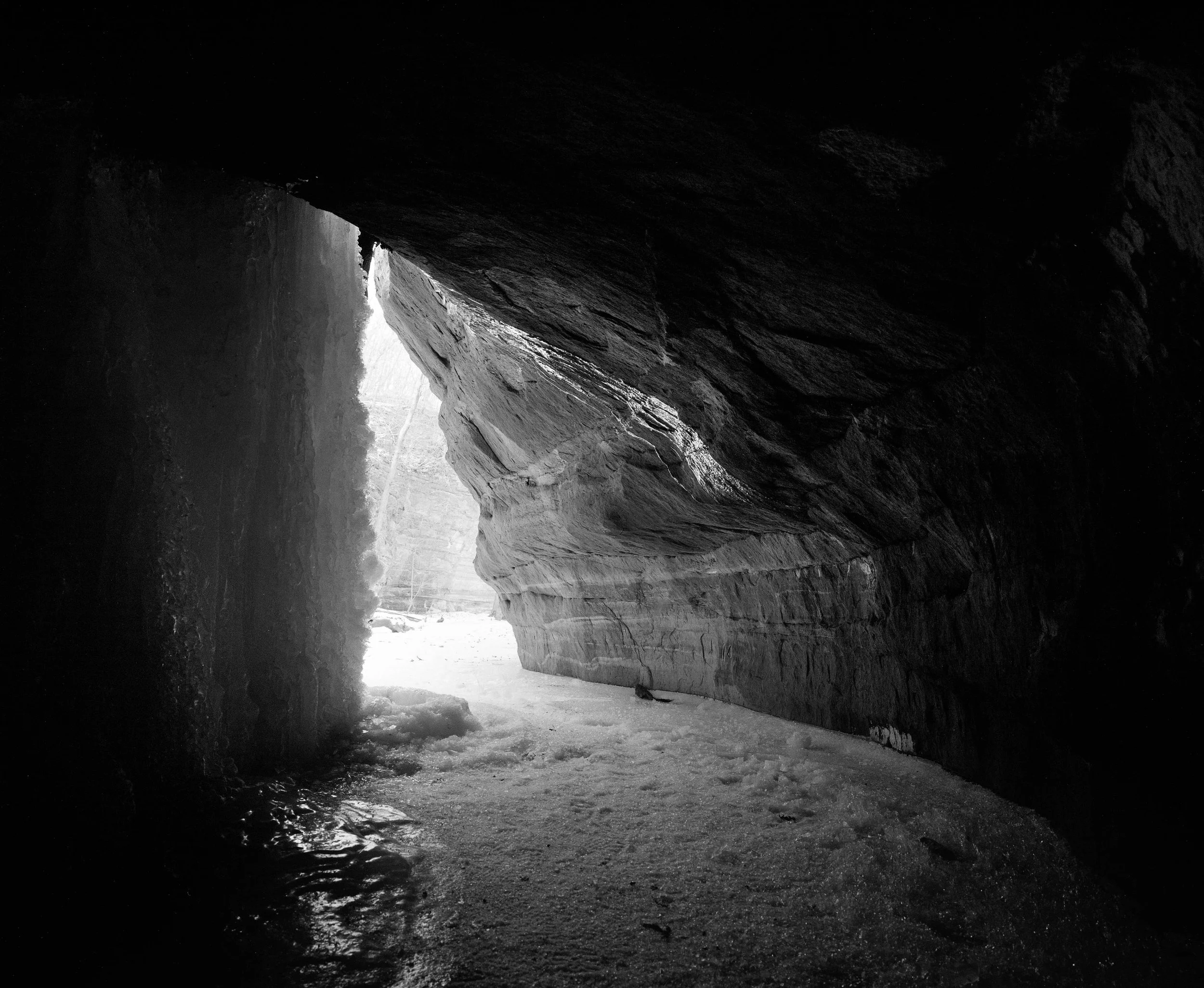 View from inside a cave looking outward, with ice and snow on the ground and a glimpse of trees and snow outside.