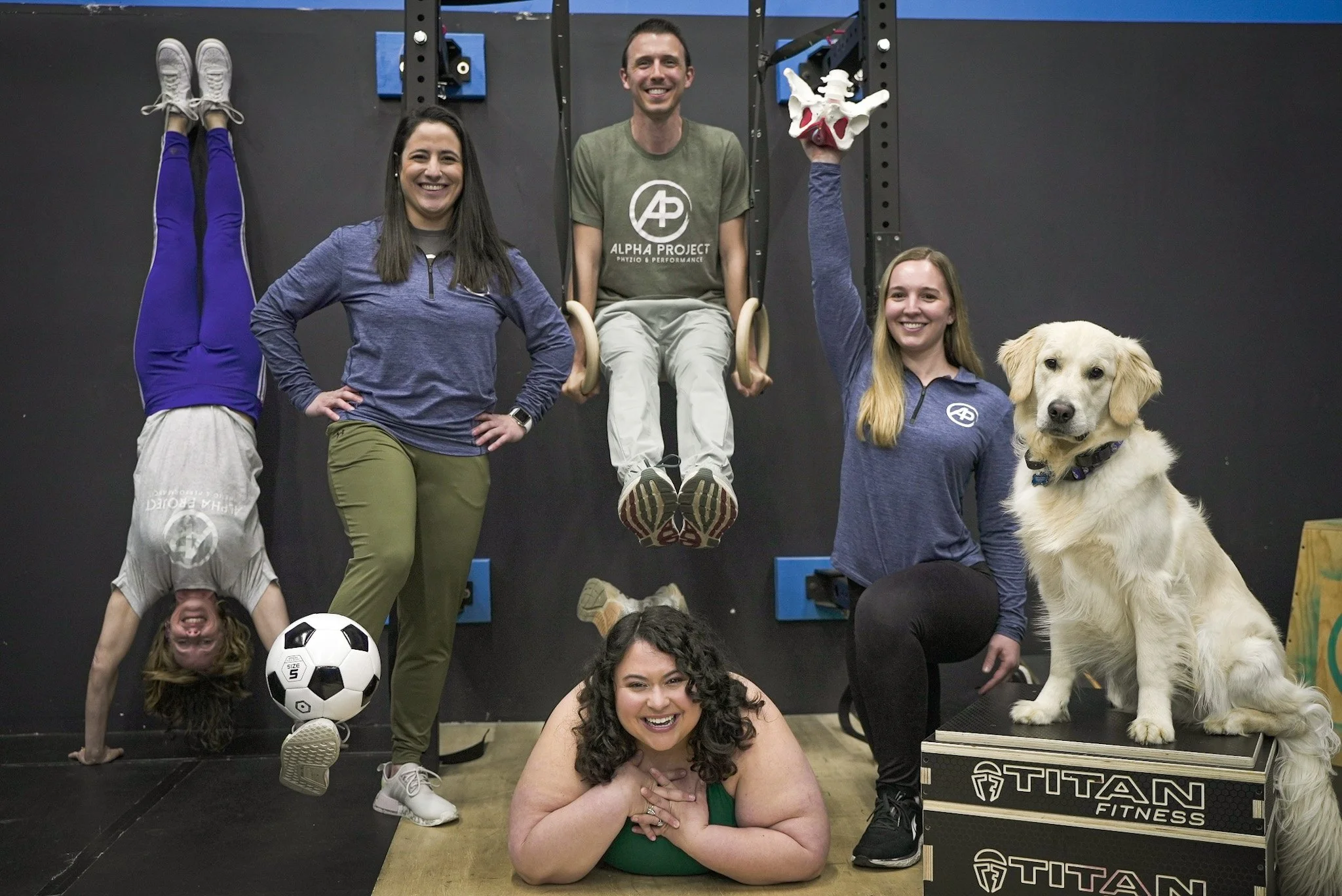 Group of people and a dog in a gym, one person doing a handstand, another holding a pelvic model, others posing with exercise equipment, dog sitting on a Titan Fitness box.