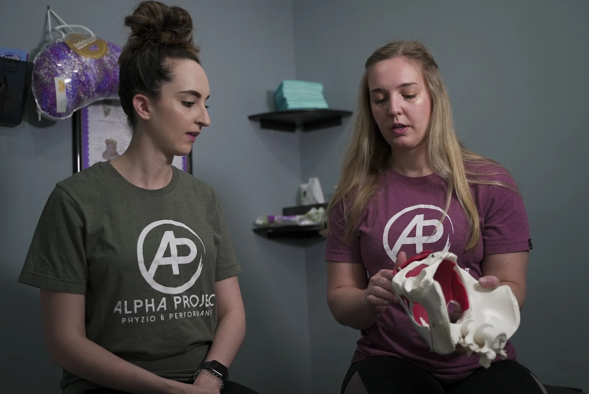 Two women wearing Alpha Project shirts, sitting in a room with shelves, discussing and holding a pelvic model.