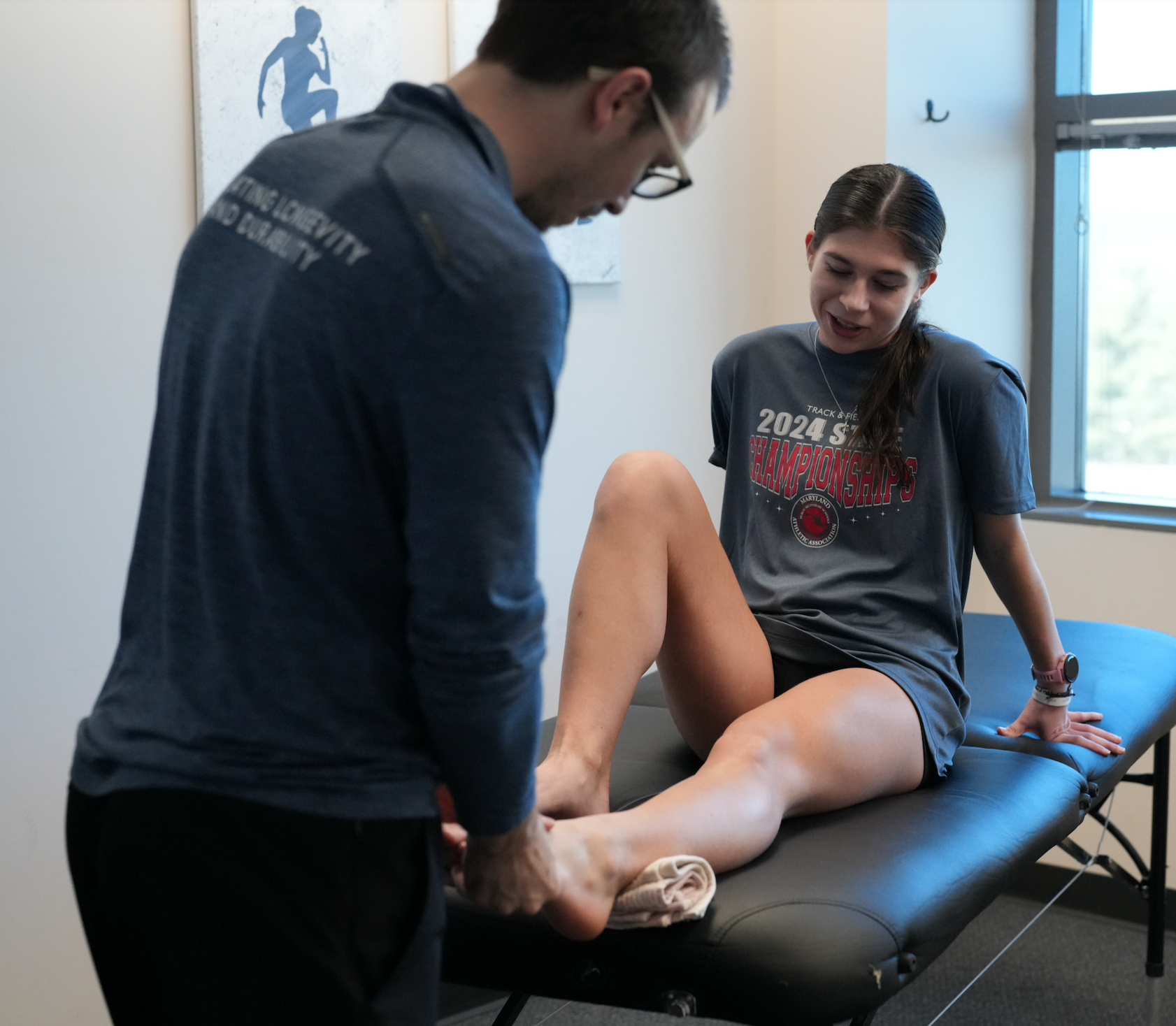 A young woman undergoing physical therapy, sitting on a treatment table with her leg raised, while a therapist supports her leg and checks her knee.