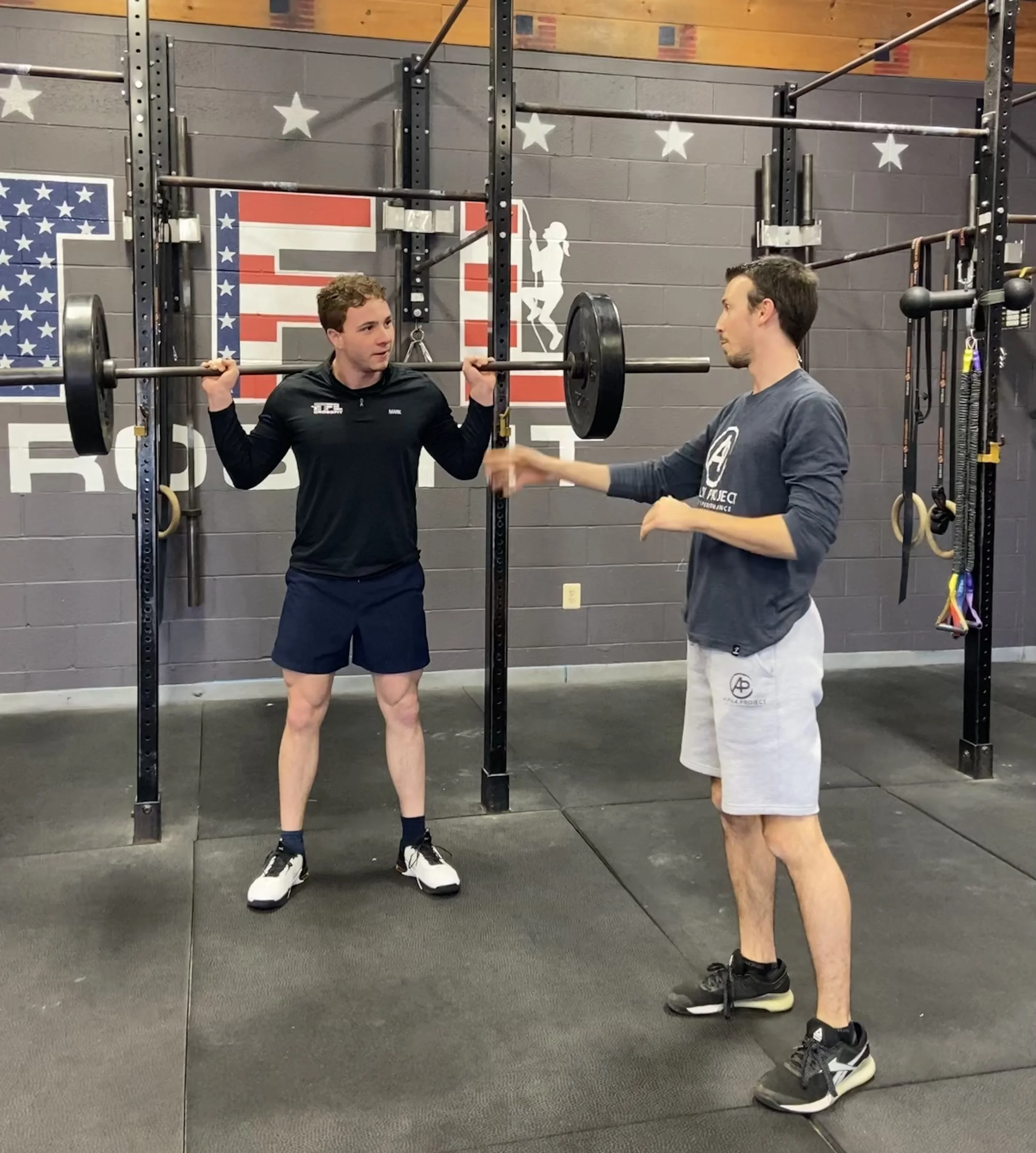 Two people in a gym, one holding a barbell on their shoulders, the other gesturing, against a wall with American flag design.