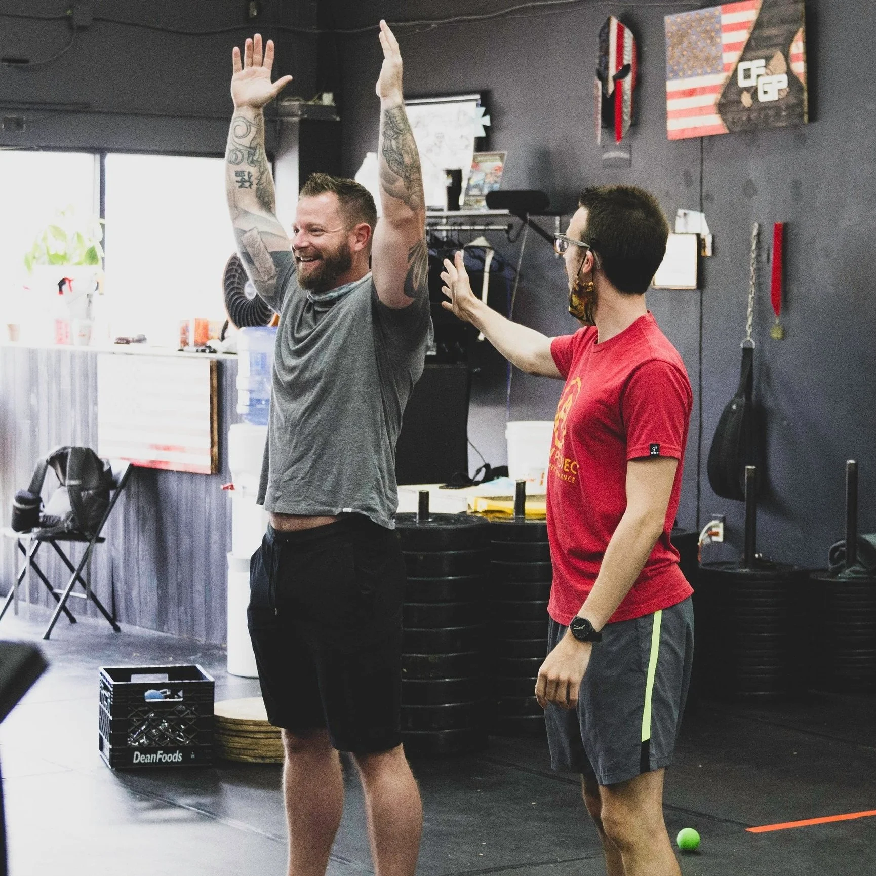 Two men in a gym, one wearing a gray shirt with tattoos and raising arms, the other in a red shirt gesturing towards him, gym equipment in the background.