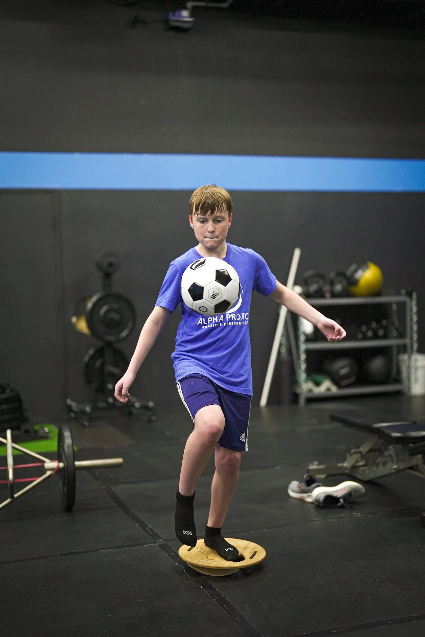 Boy balancing on a wobble board while kicking a soccer ball in a gym setting.