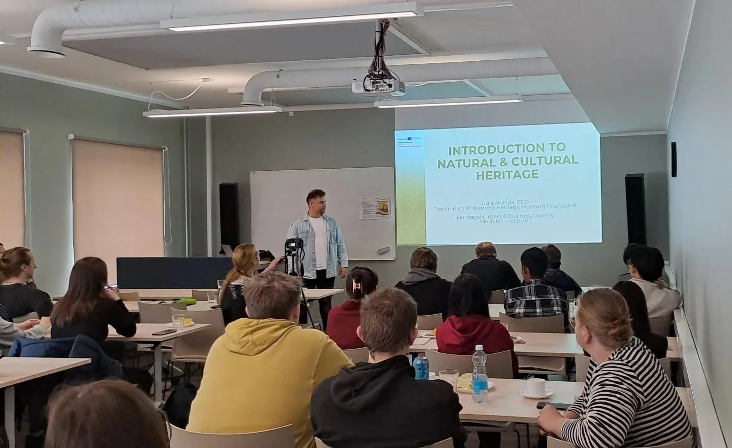 Kuva luokkahuoneen takaosasta. Etupulpetit täynnä innokkaita opiskelijoita ja edessä seisoo luennoitsija.