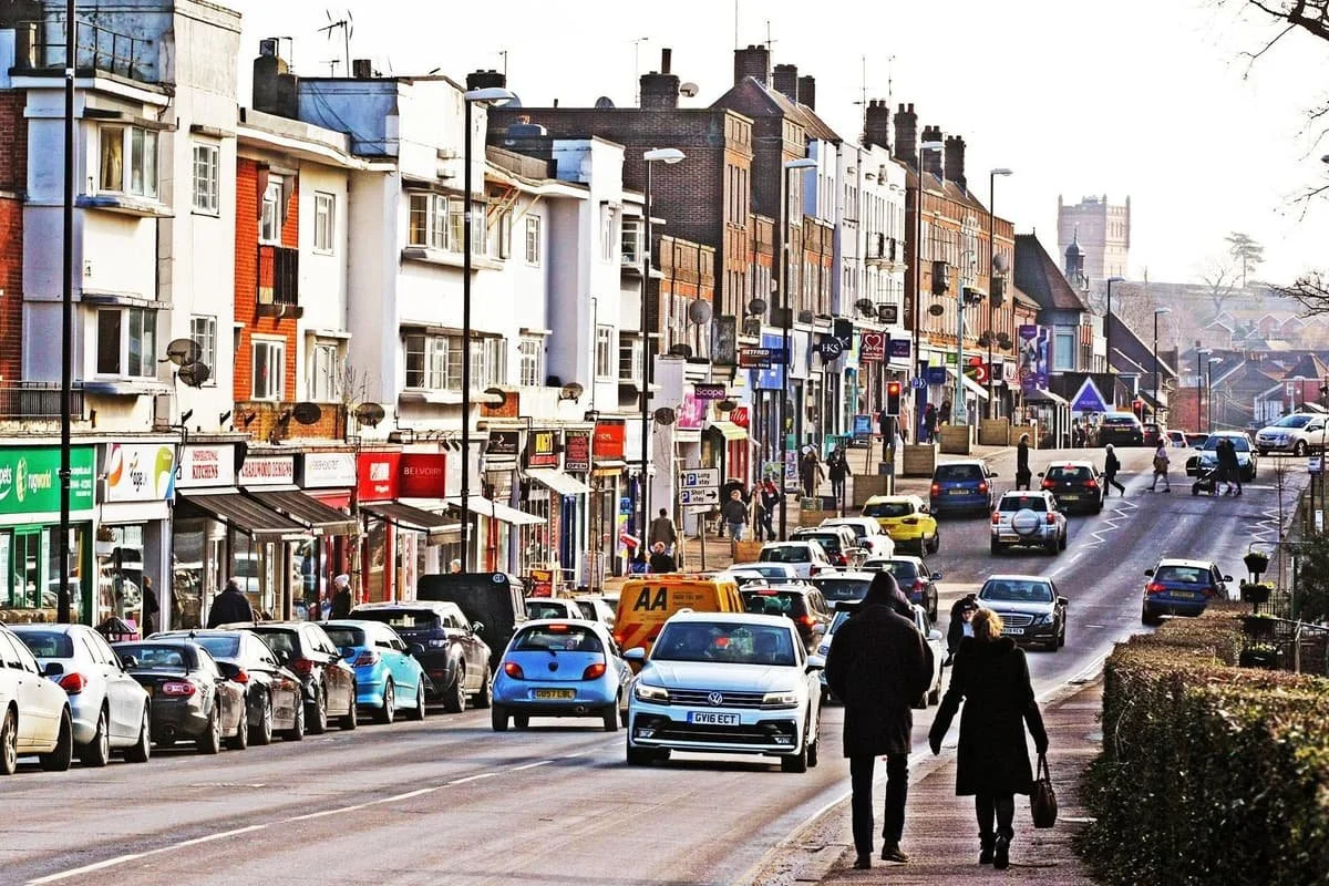 City street scene with parked and moving cars, pedestrians walking on the sidewalk, buildings with various storefronts, and a distant church tower.