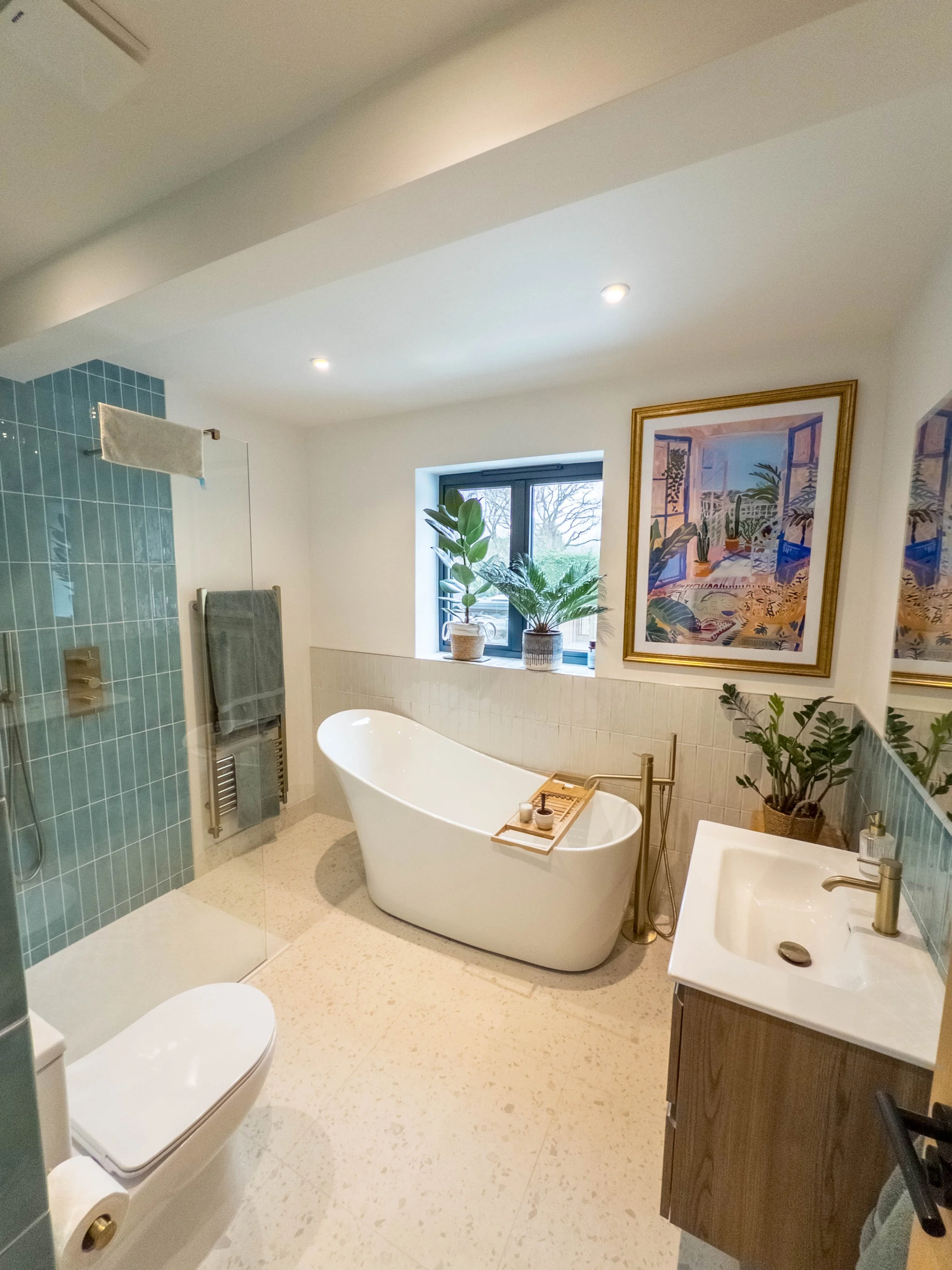 Modern bathroom with a freestanding bathtub, a window with potted plants, a framed artwork, a glass-enclosed shower, a toilet, and a sink with greenery.