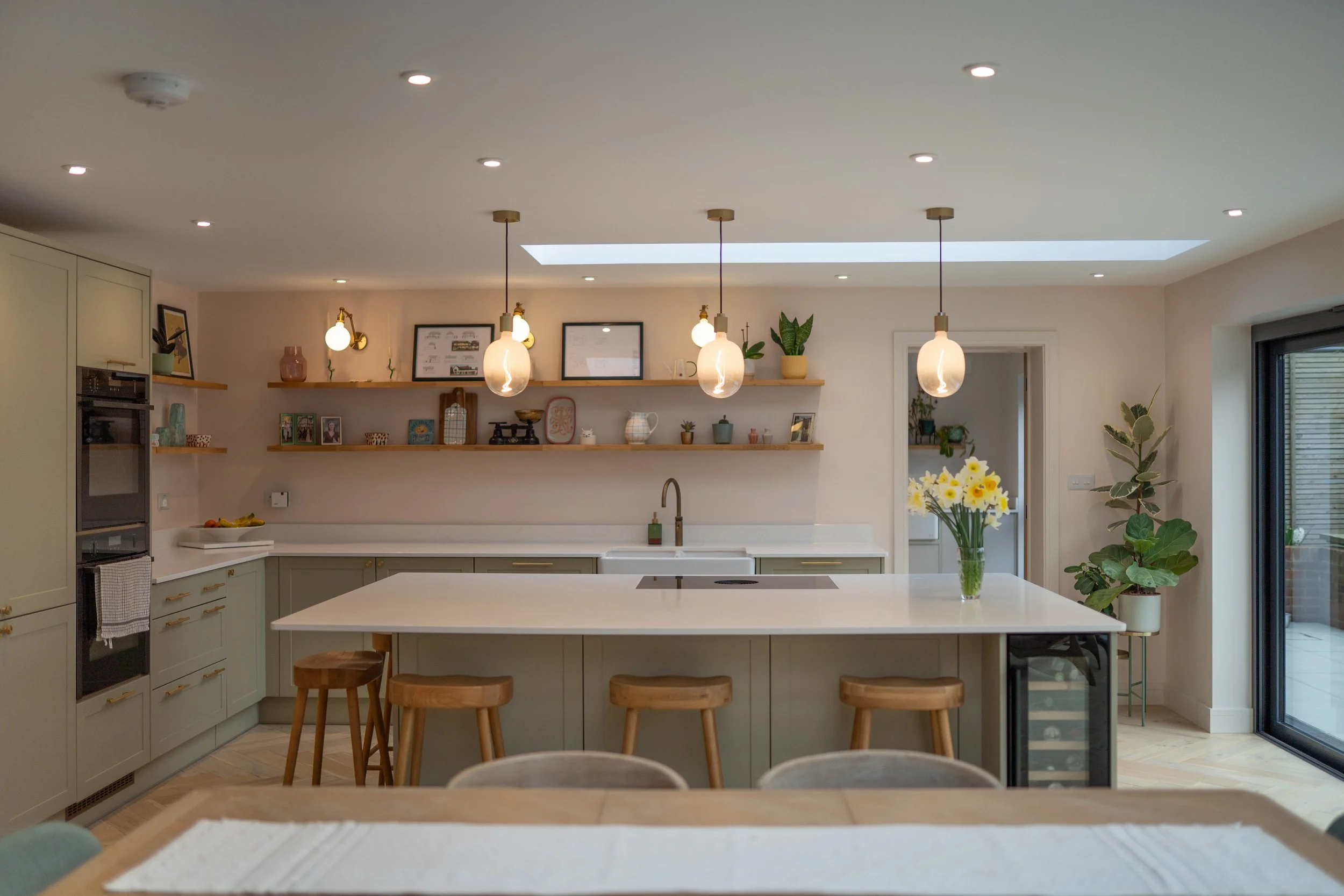 Modern kitchen with light green cabinets, white island with wooden stools, pendant lights, open shelf with decor, and large sliding glass door to outdoor patio.