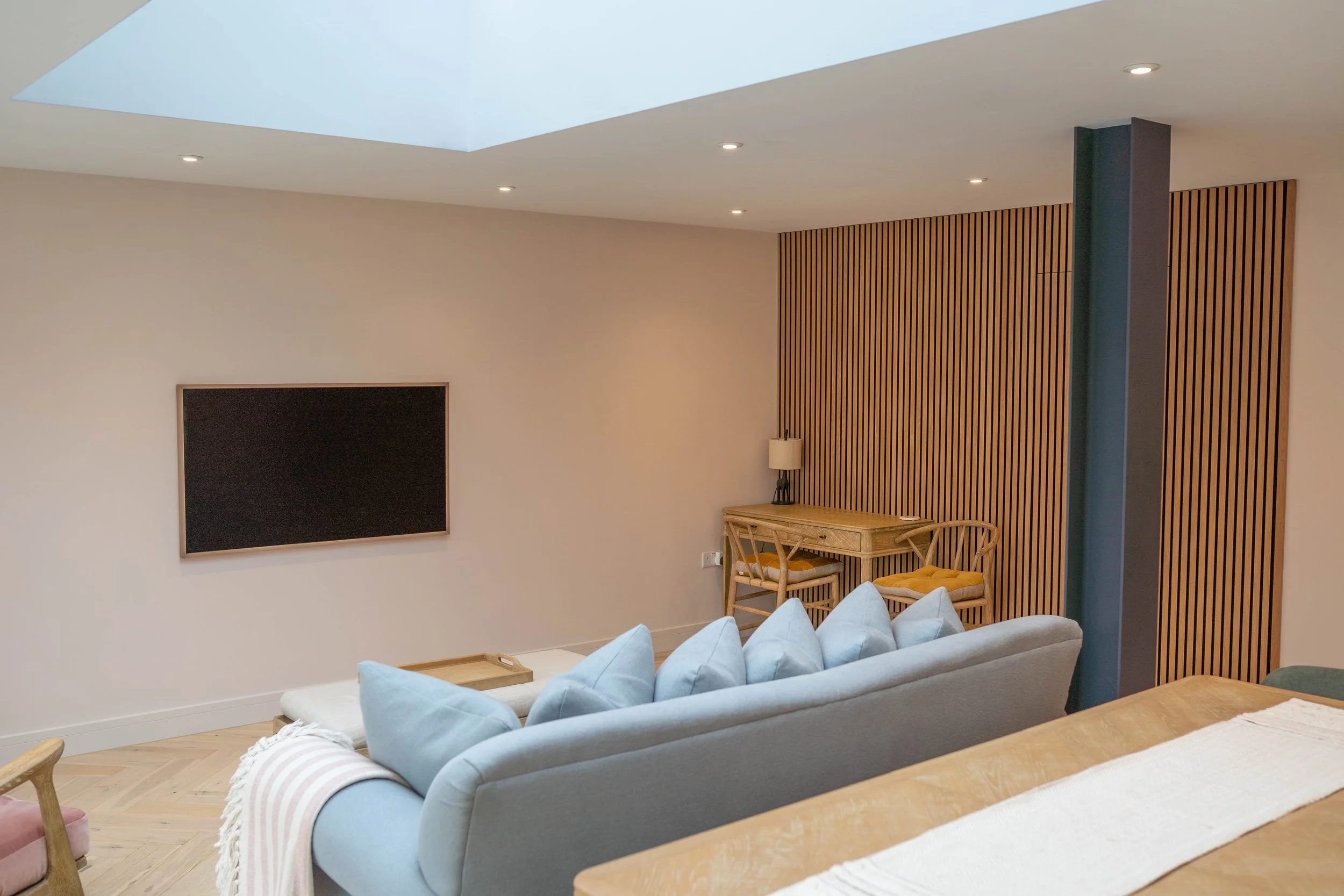 Modern living room with a TV on a beige wall, a light gray sofa with blue pillows, a wooden table, and a small desk with chairs against a wooden slatted wall. Skylight above, recessed ceiling lights.