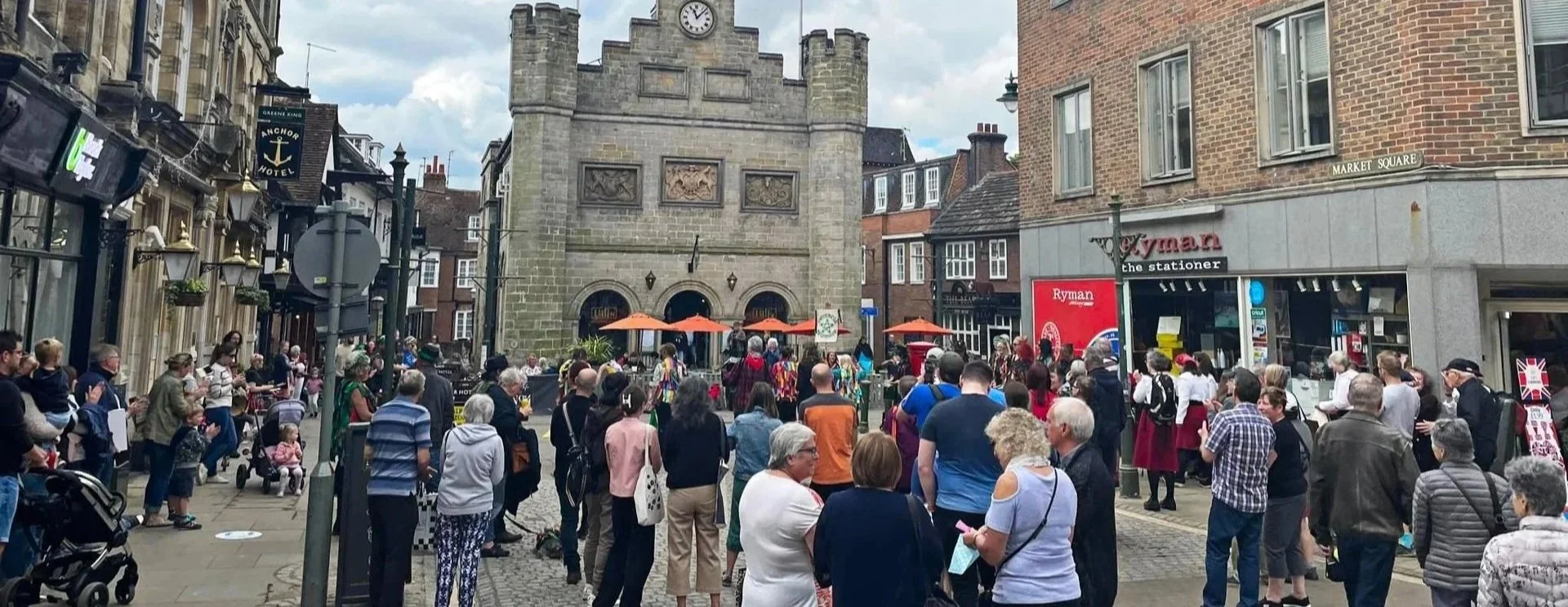 Crowd gathered in a town square with historic buildings, a castle tower, and a performance by people in traditional costumes, with some spectators taking photos.