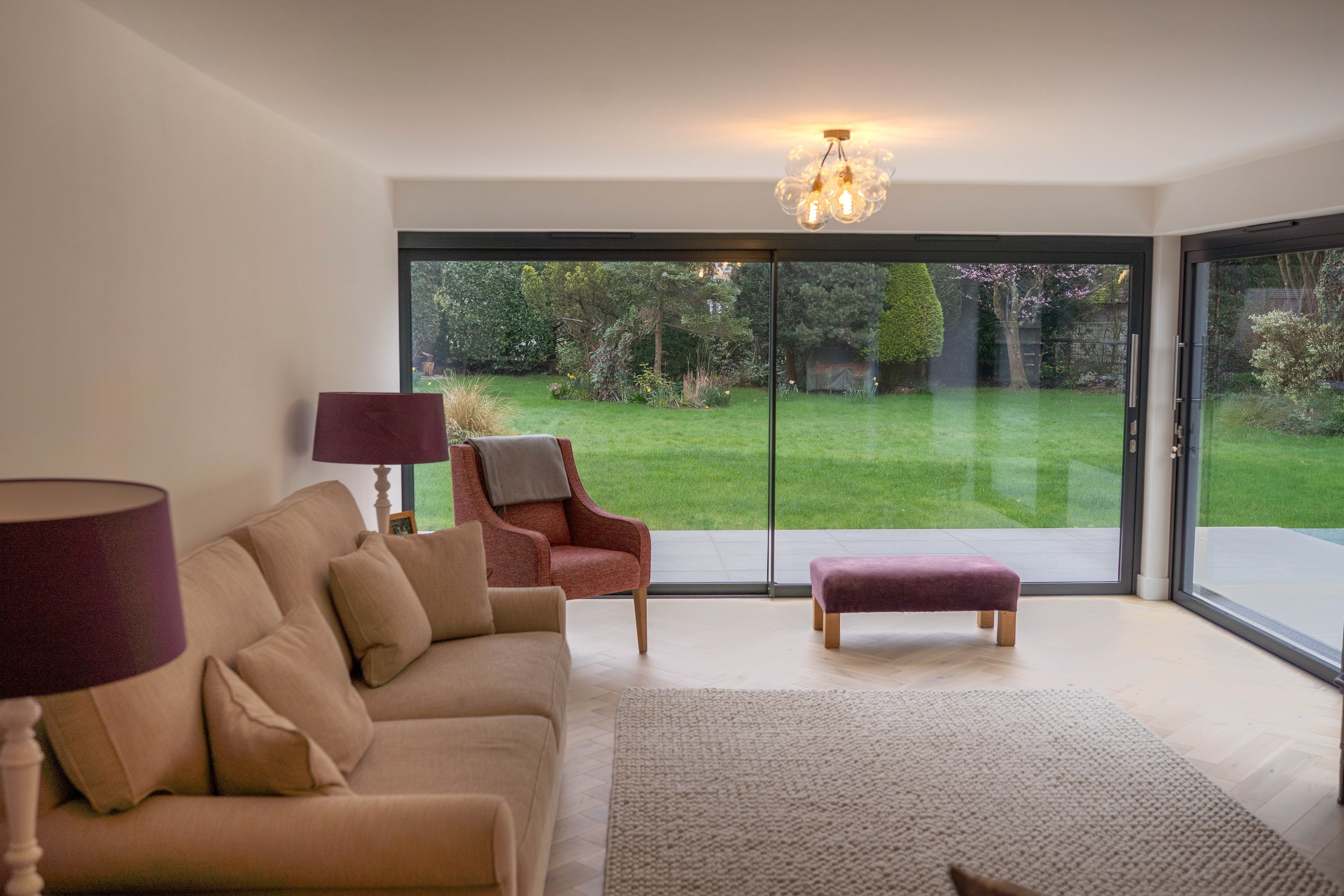 Interior of a living room with a beige sofa, purple lamps, a red armchair with a blanket, a purple ottoman, and large sliding glass doors leading to a green backyard.