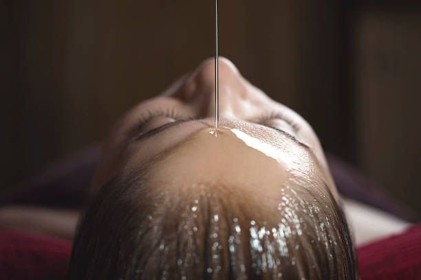 Une personne allongée, recevant un traitement facial ou capillaire avec un liquide versé sur son front, photographié de dessus.