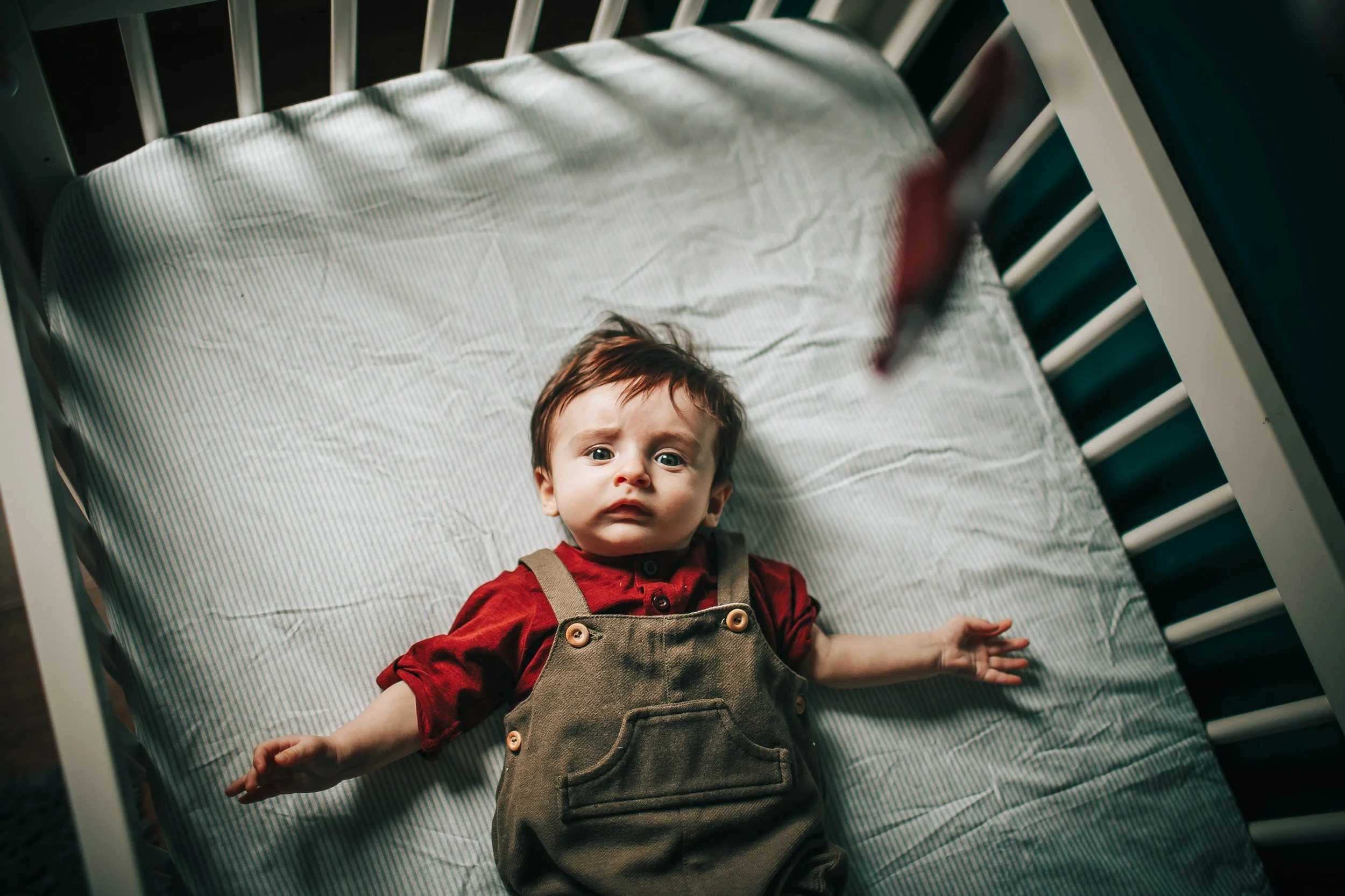 A baby lying in a crib, wearing a red shirt and khaki overalls, looking up with a curious expression.