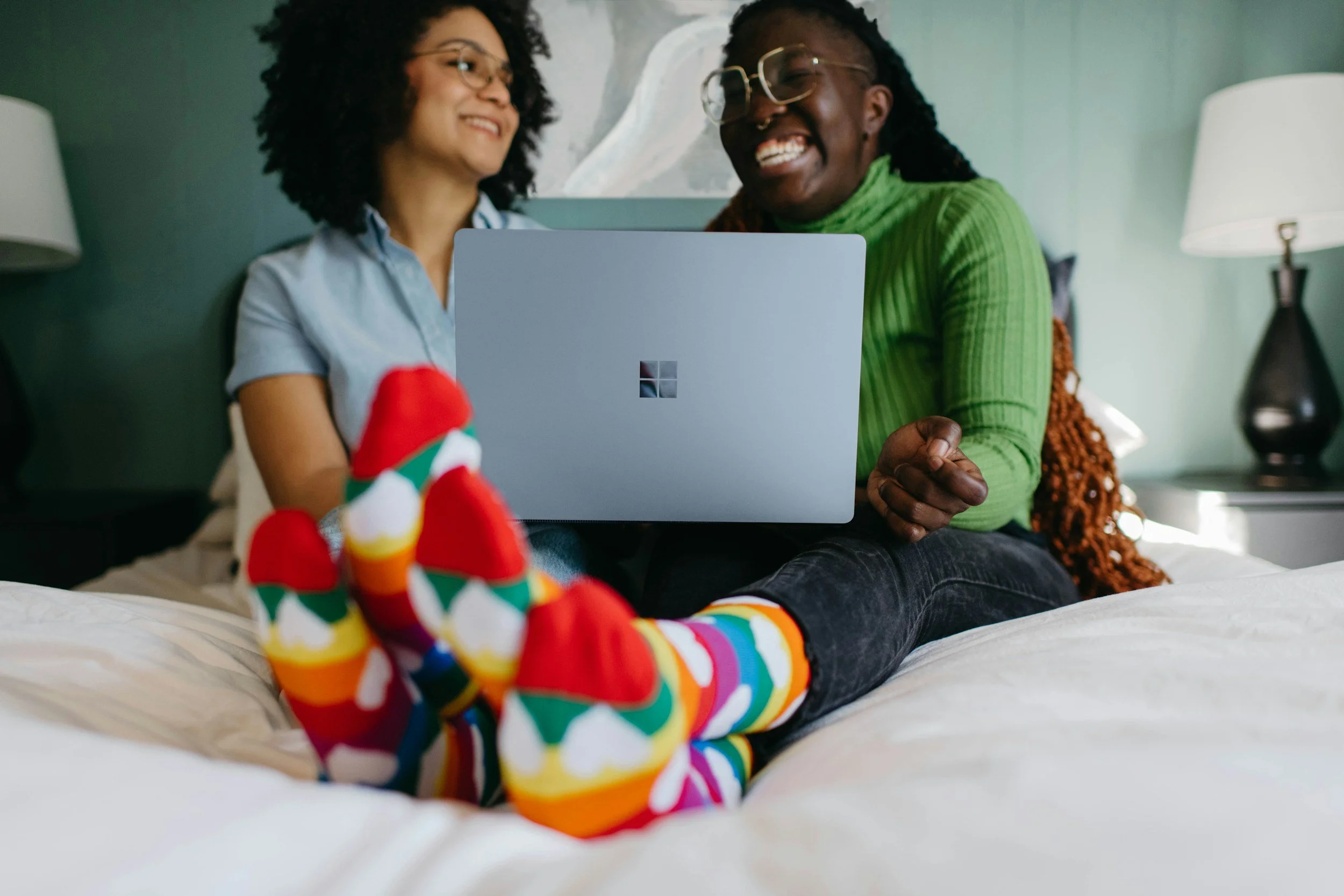 Two women smiling and sitting on a bed, looking at a Microsoft surface laptop. One woman is wearing colorful rainbow socks and a bright green sweater, the other is wearing glasses and a light blue shirt. There are nightstands and lamps in the background.