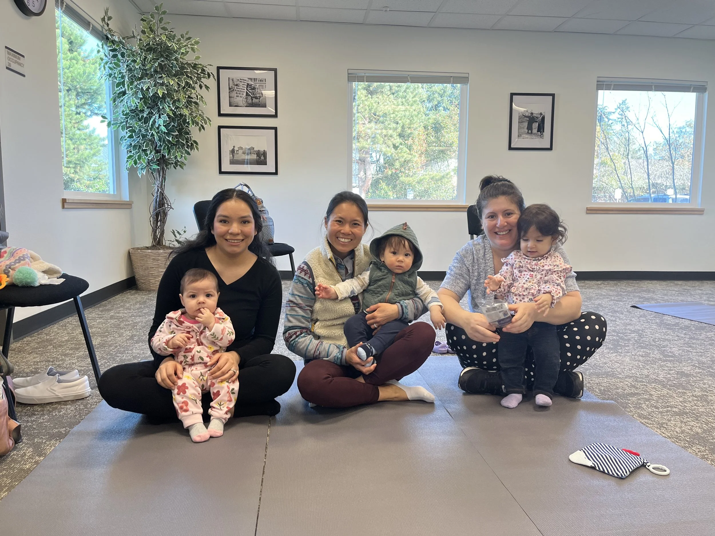 women and four children sitting on the floor of a room with large windows and framed black-and-white photographs on the wall. The women are smiling, and the children are sitting on their laps or beside them. There are plants and chairs in the room, and personal belongings such as shoes and a small bag are visible.