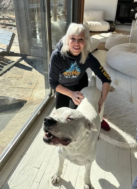 Dr. Lila Wolfe, animal chiropractor, a woman with blonde hair smiling and treating a large white dog inside a modern living room, with large windows and a patio visible outside.