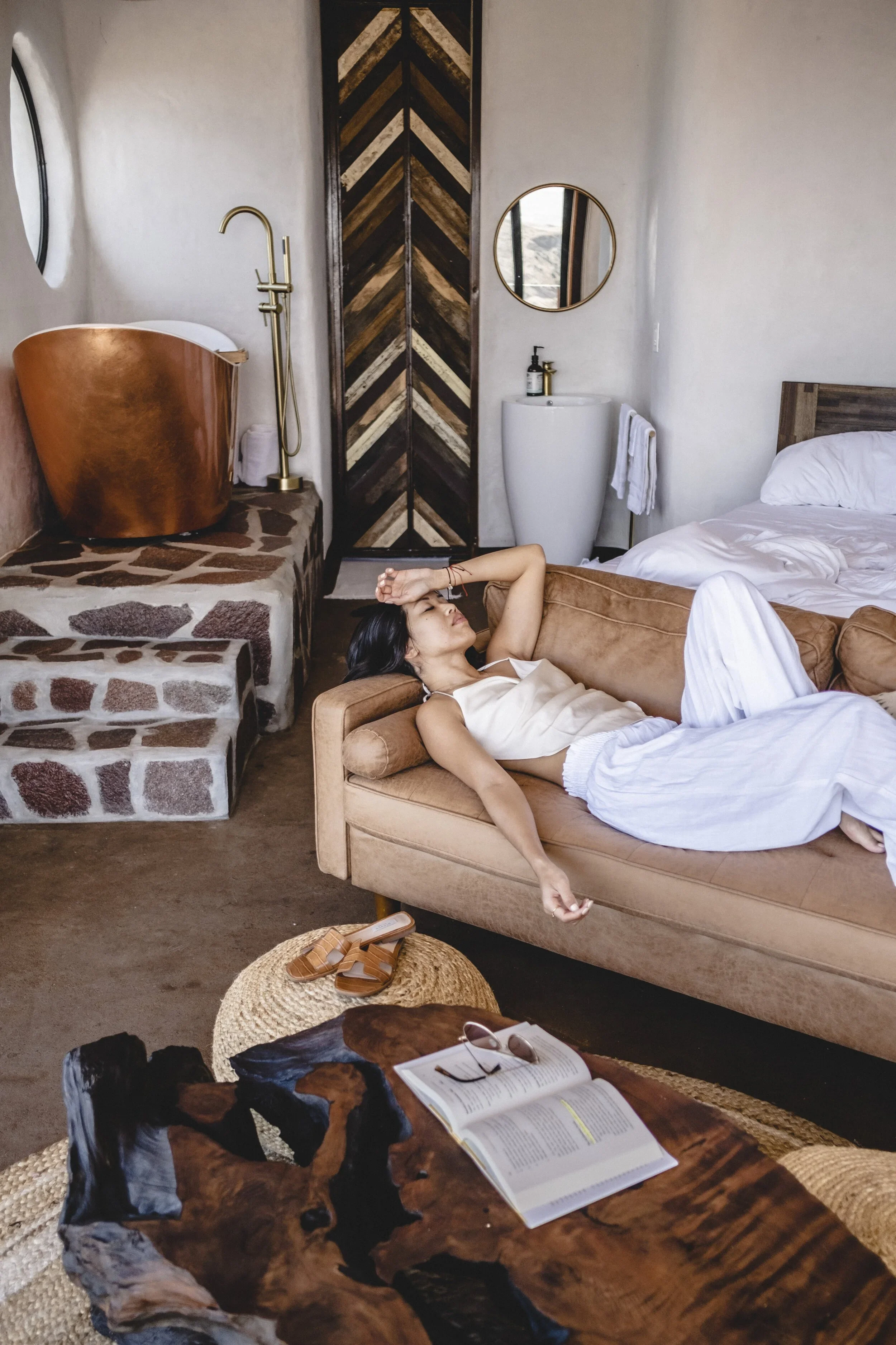 Person lying on a beige sofa with white pillows, resting in a room with rustic decor including a round mirror, a wooden door with chevron pattern, a copper bathtub, and a small vanity with a round mirror, book, sunglasses, and a sculpture of a dog on a wooden table, with a woven basket containing sandals in the foreground.