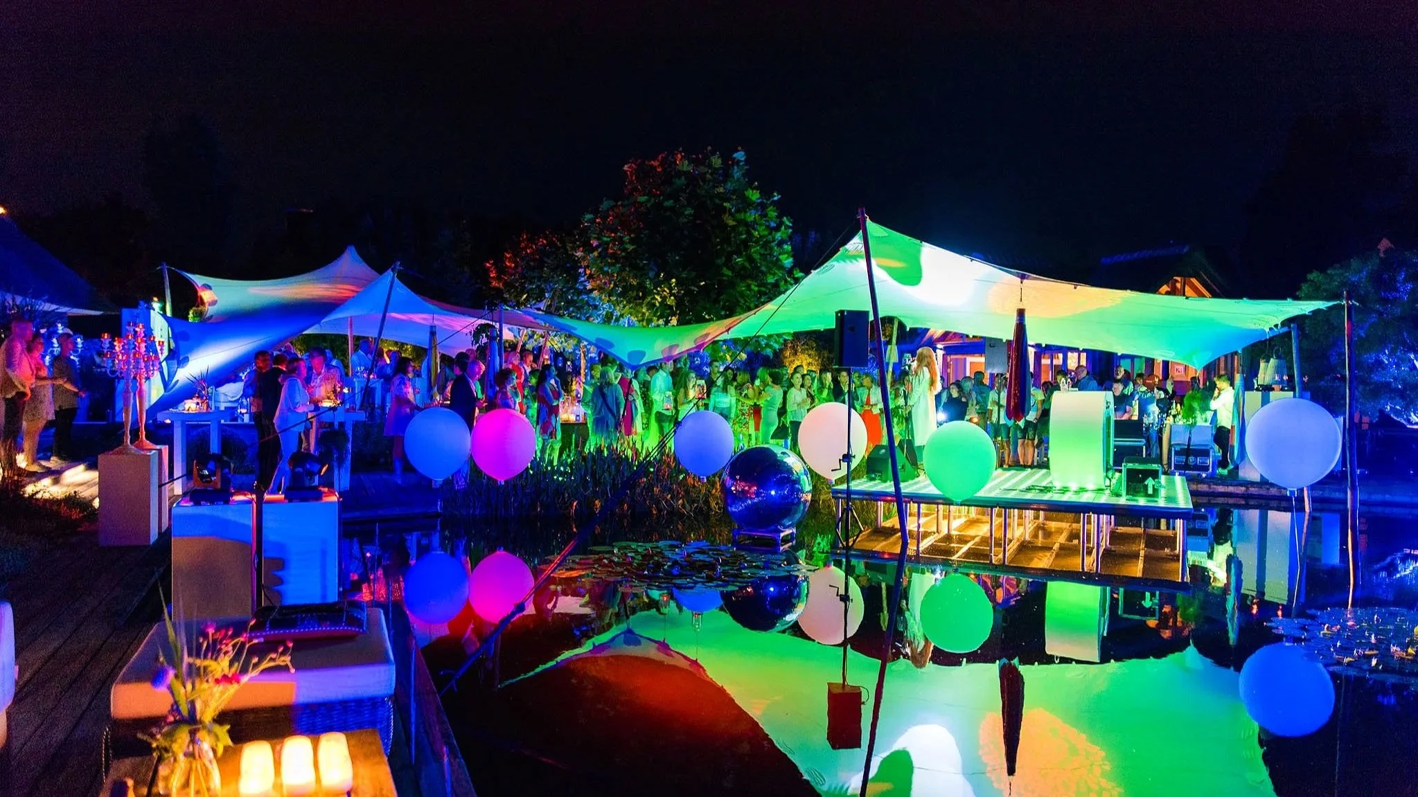 Nachtelijk buitenfeest met kleurrijke verlichting, tenten en ballonnen. Mensen verzamelen zich onder de tenten naast een verlichte vijver.