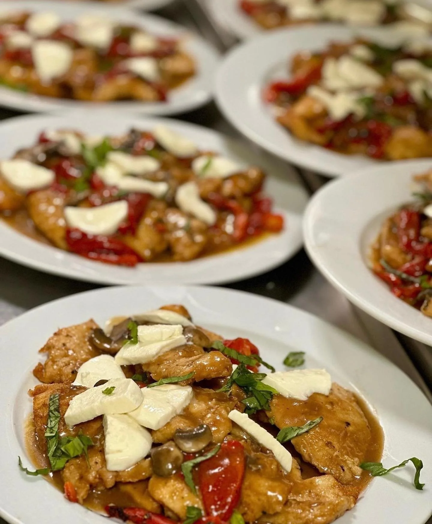 Several white plates filled with chicken breast topped with creamy white cheese, along with sliced mushrooms, red bell peppers, and chopped green herbs.