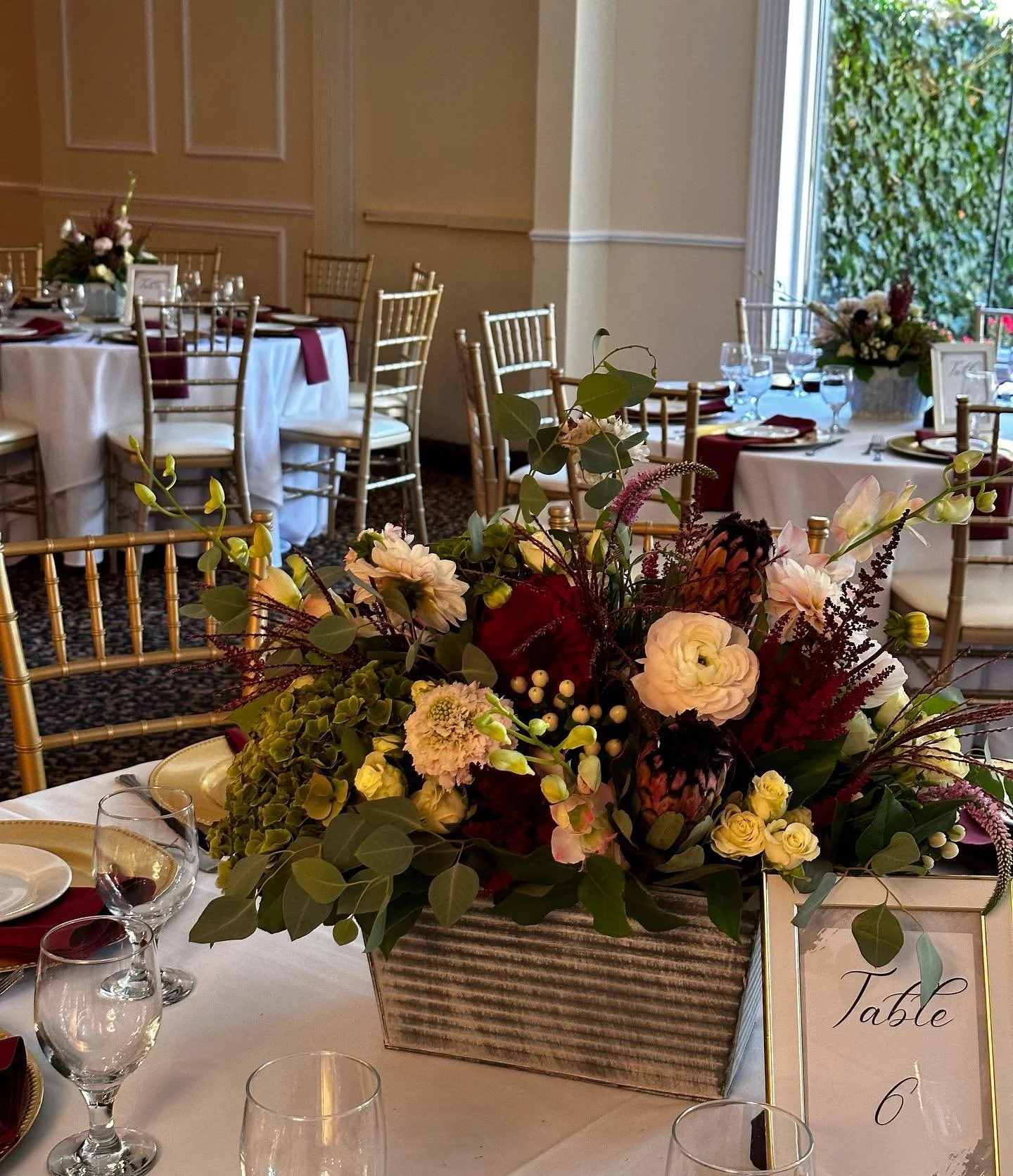 A floral centerpiece with various flowers and greenery on a table at an event. Multiple tables with chairs and place settings are visible in the background, with a window and greenery outside.