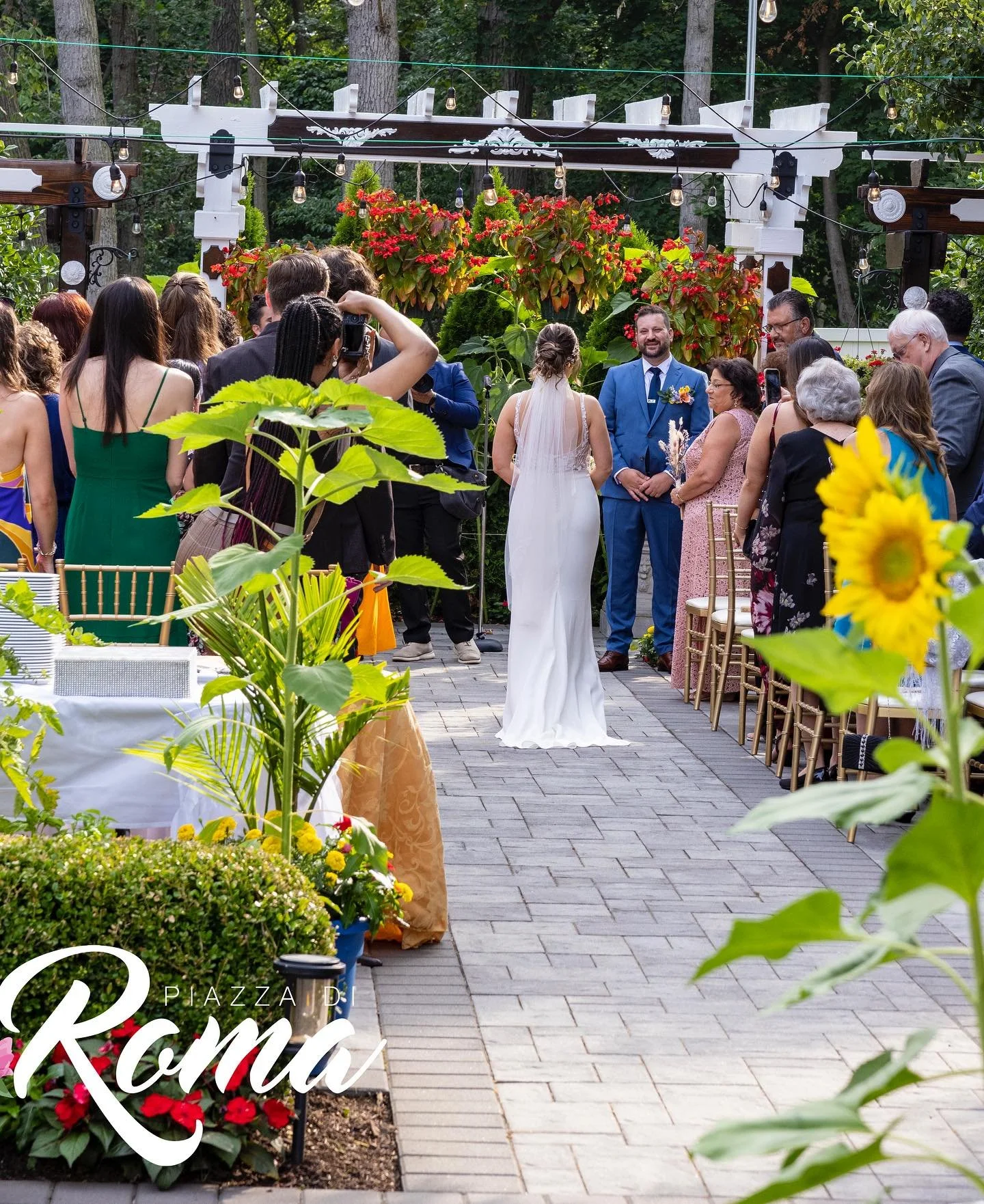 A wedding ceremony taking place outdoors in a garden setting with trees in the background. The bride in a white gown and veil stands facing the groom in a blue suit under a decorative arch. Guests are gathered around, some taking photos, seated and s