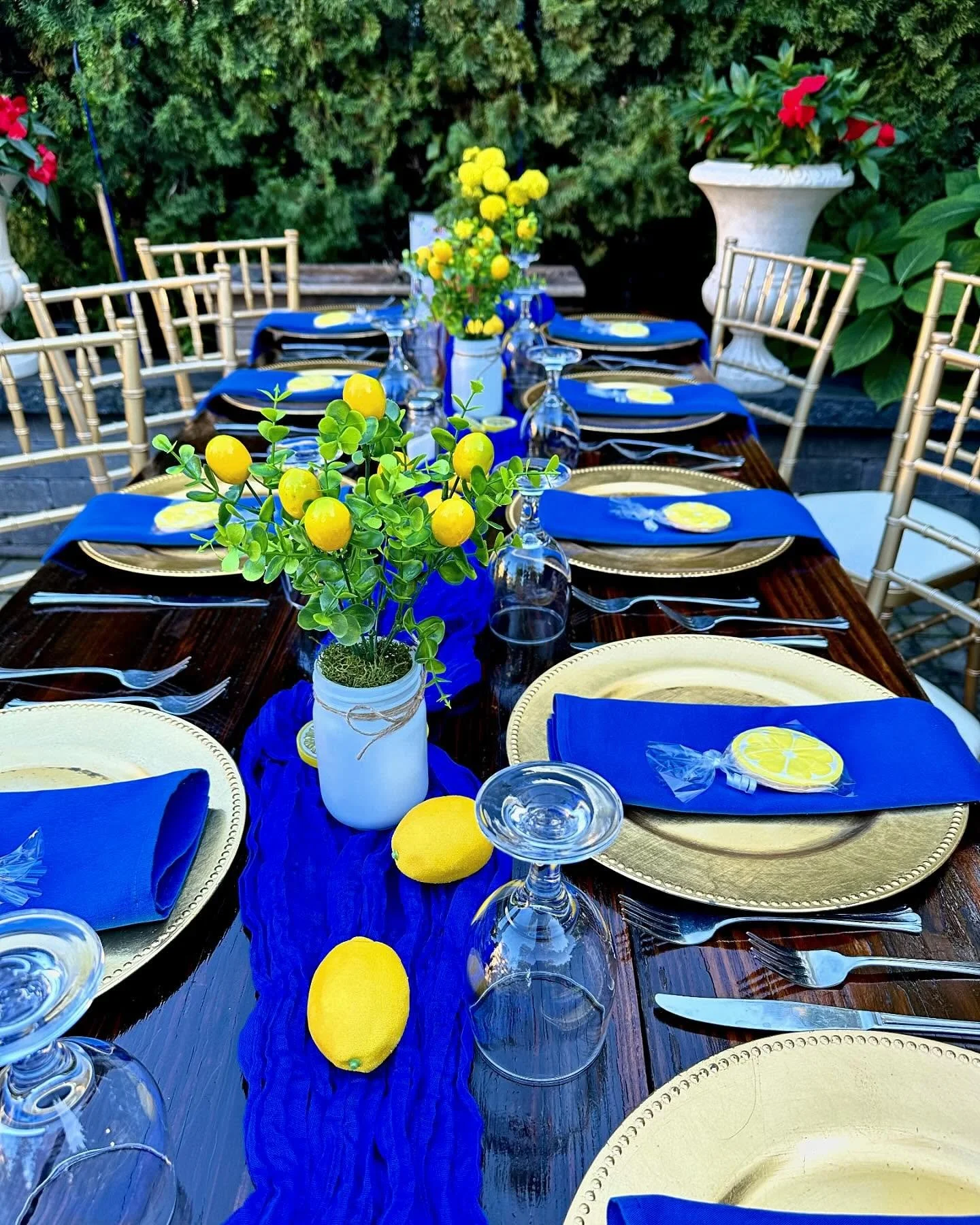 Table set outdoors with gold charger plates, blue napkins, clear glasses, and lemon-themed decorations. Yellow lemons and a lemon-themed centerpiece are arranged on a dark wooden table, with a blue table runner, surrounded by gold chairs and potted p