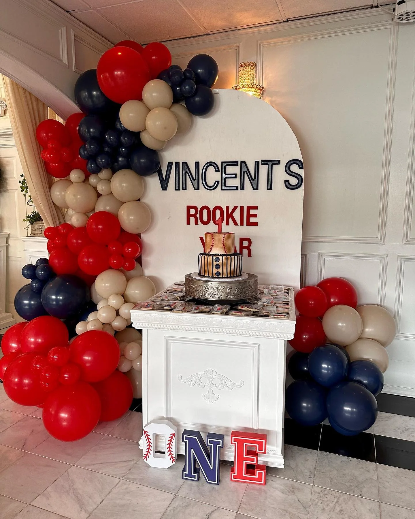Decorations for a first birthday celebration, featuring a white backdrop with the name 'VINCENT' and the words 'ROOKIE YEAR' in red and black lettering, surrounded by red, white, and navy blue balloons, and a cake on a white table with colorful decorations.