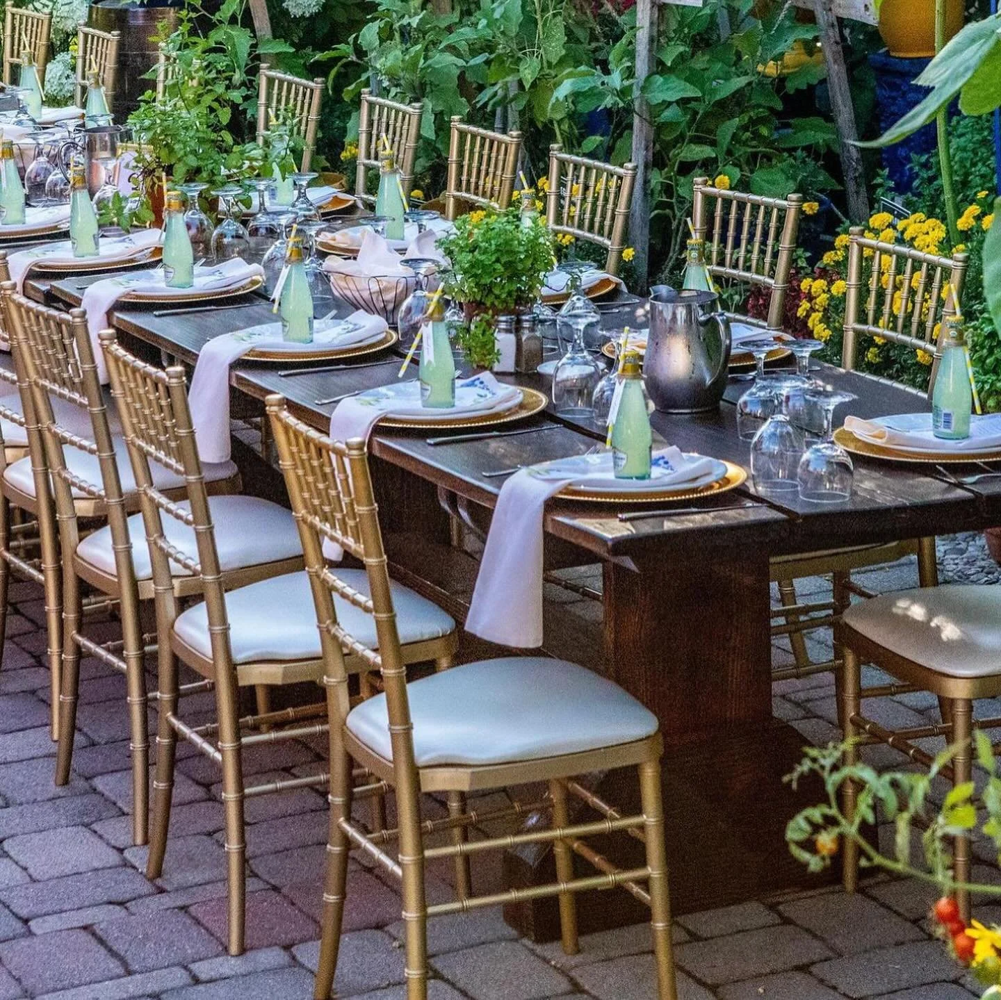 An outdoor dining table set with gold chairs, white napkins, plates, glassware, and water bottles on a brick patio surrounded by lush green plants and yellow flowers.