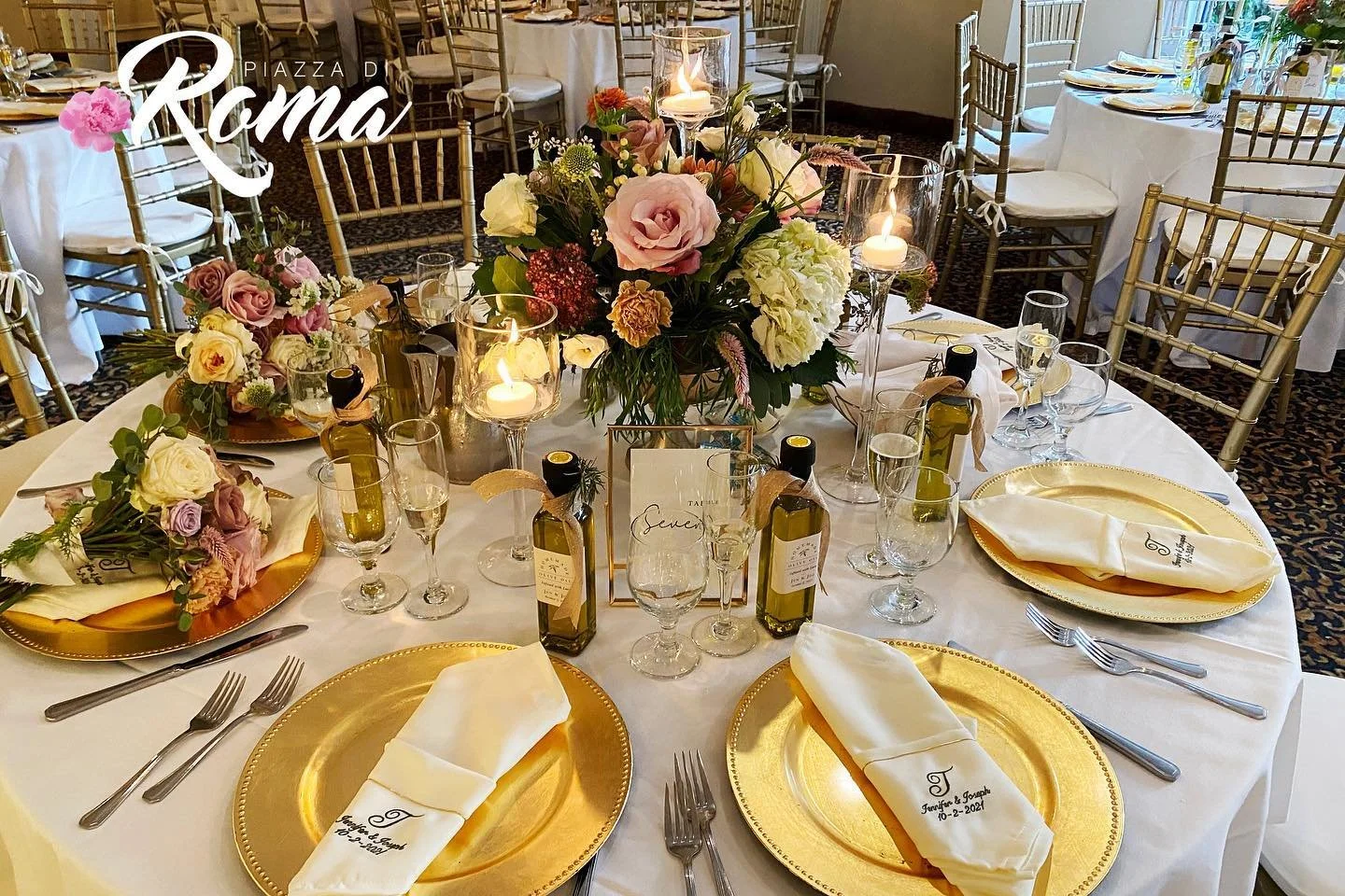 A round wedding reception table decorated with gold chargers, white napkins embroidered with the couple's names and wedding date, floral centerpieces with pink, white, and purple flowers, surrounded by gold and silver flatware, wine glasses, and small bottles of olive oil, in a ballroom with gold Chiavari chairs and white tablecloths.