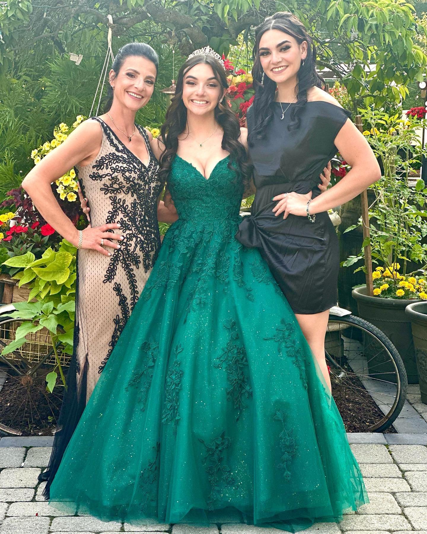 Three women standing outdoors among greenery, dressed in formal attire. The woman in the middle wears a green ball gown with floral embroidery, and a tiara. The woman on the left wears a beige dress with black lace details, and the woman on the right wears a black dress.