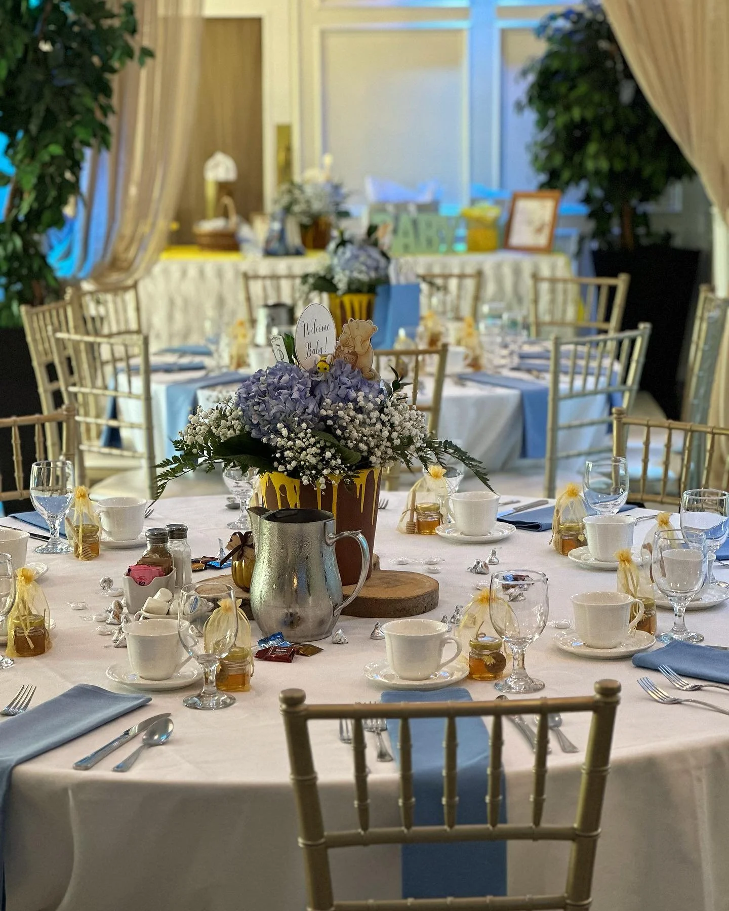 A decorated banquet table set for a celebration with a floral centerpiece, teacups, glasses, silverware, and small gift bags, in a room with elegant curtains and additional tables in the background.