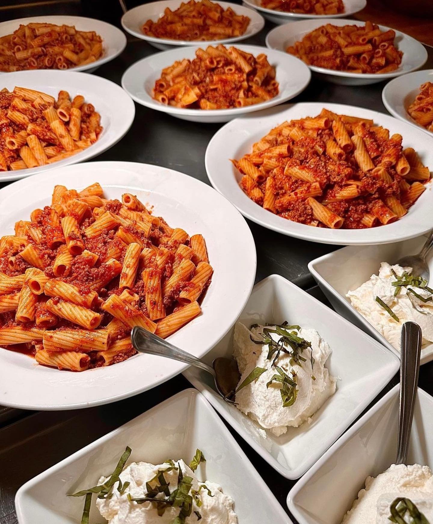 Several plates of cooked pasta with red sauce and bowls of white creamy cheese or whipped topping garnished with herbs.