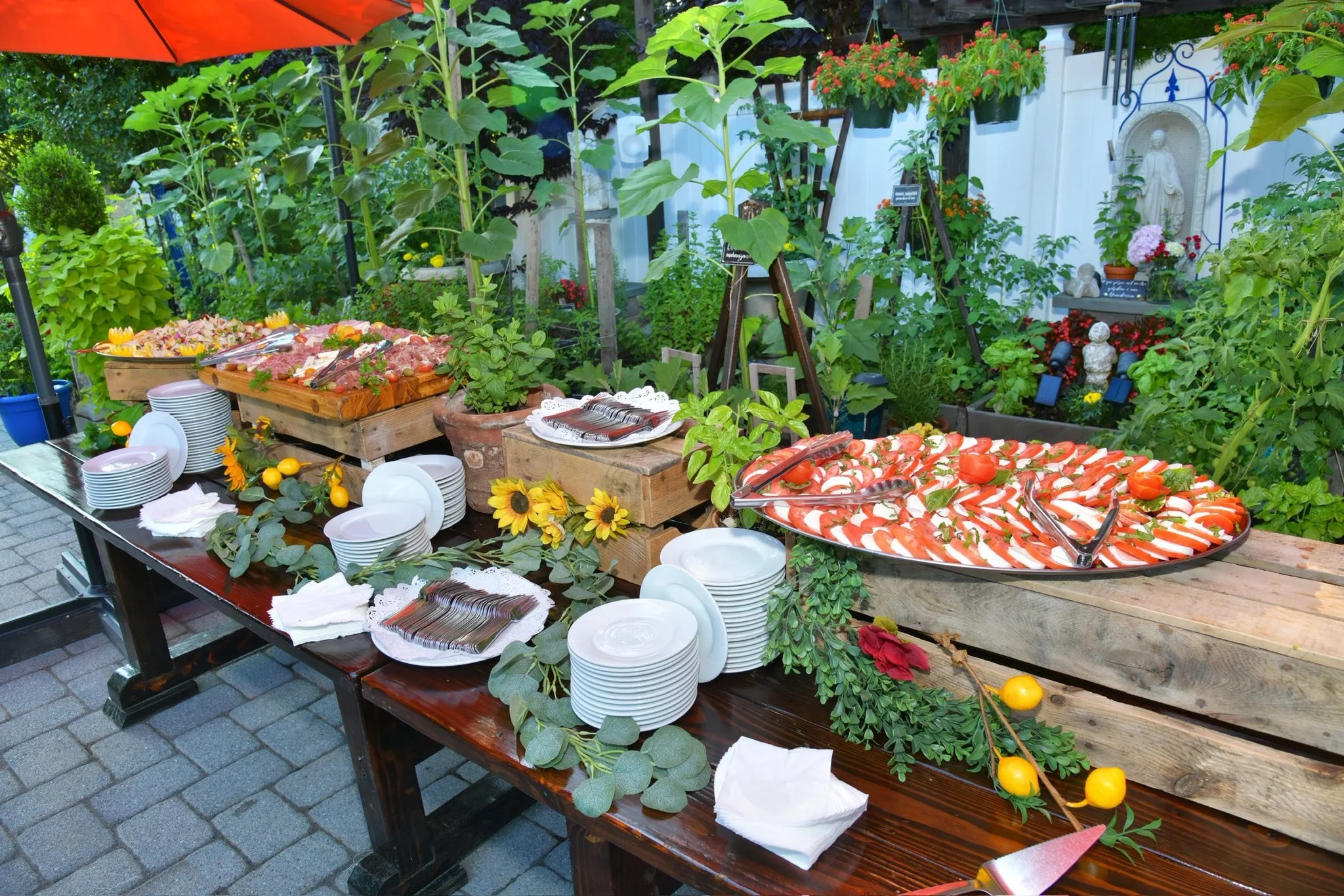 Outdoor buffet table with slices of tomatoes and mozzarella, trays of sliced meats, plates, utensils, and decorative yellow flowers in a garden setting with green plants and religious statues in the background.