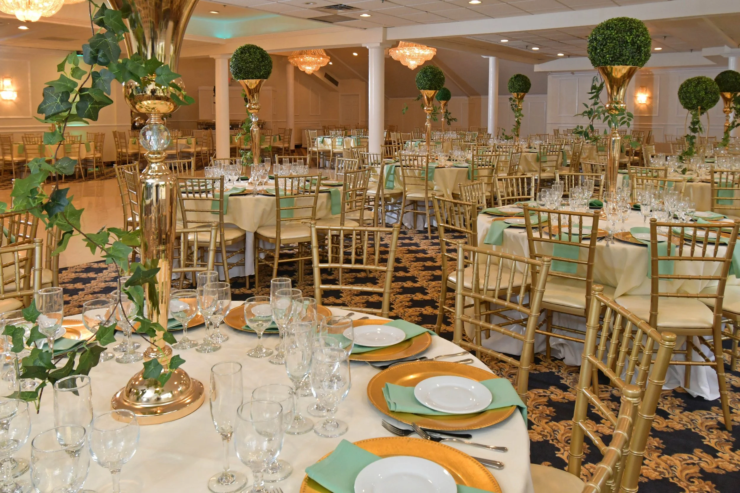 Elegant banquet hall decorated for a formal event with round tables, gold chairs, and tall floral centerpieces in gold vases.