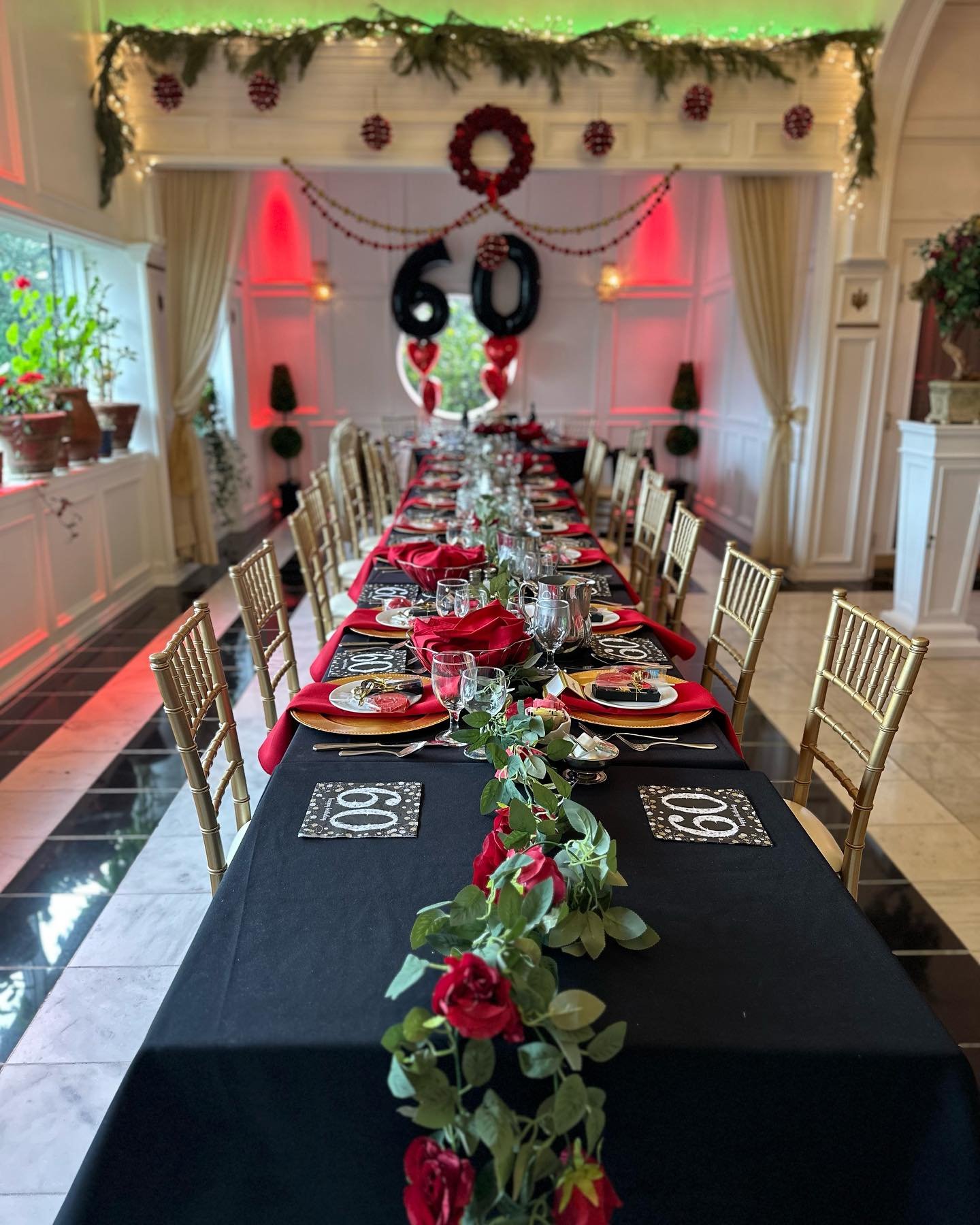 Decorated banquet table for a 60th birthday celebration with black and red theme, gold chairs, floral centerpiece, and 60 balloons on the wall