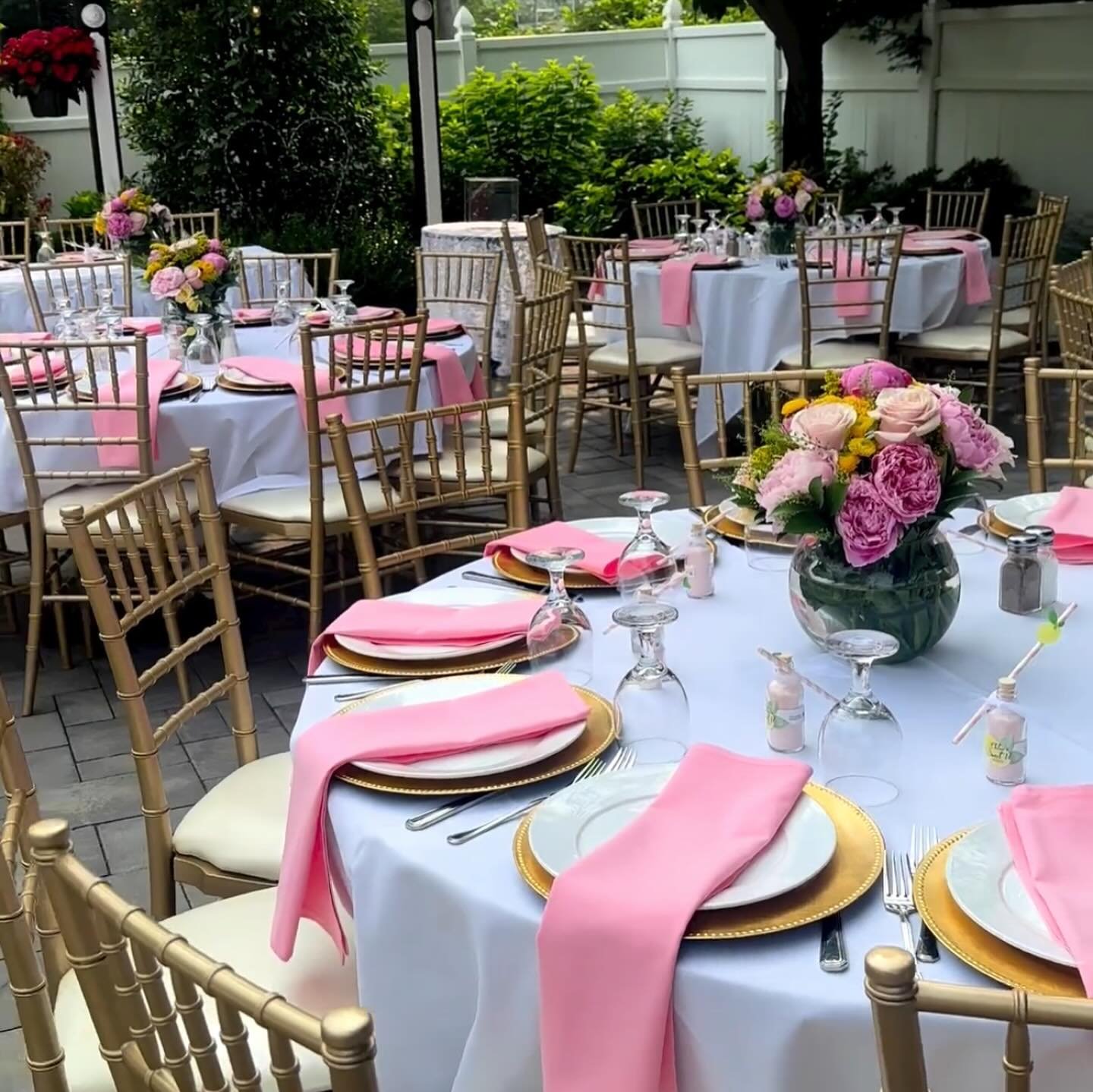 Outdoor dining area decorated for a celebration with round tables covered in white tablecloths, pink napkins, gold-rimmed plates, and floral centerpieces with pink, purple, and yellow flowers. Gold chairs surround the tables, and the setting is compl