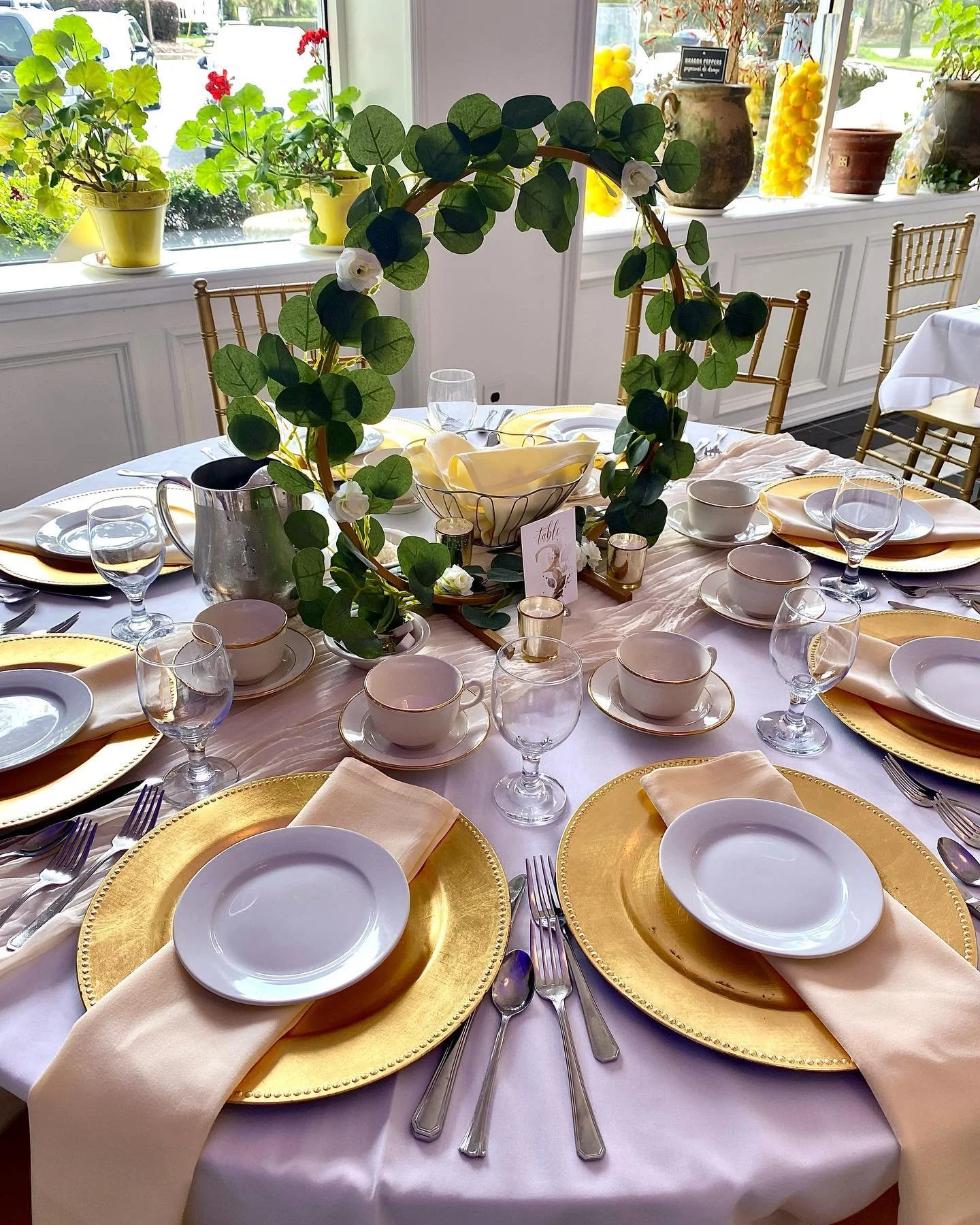 A round dining table set for a formal event with gold chargers, white plates, beige napkins, and glassware, decorated with a circular green leaf and white flower centerpiece, in a room with large windows and decorated with yellow and brown pots and p