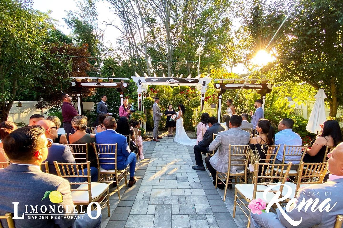 An outdoor wedding ceremony taking place in a garden with guests seated on gold chairs and the bride and groom standing under a decorated archway. The sun is shining through trees, creating a warm glow.