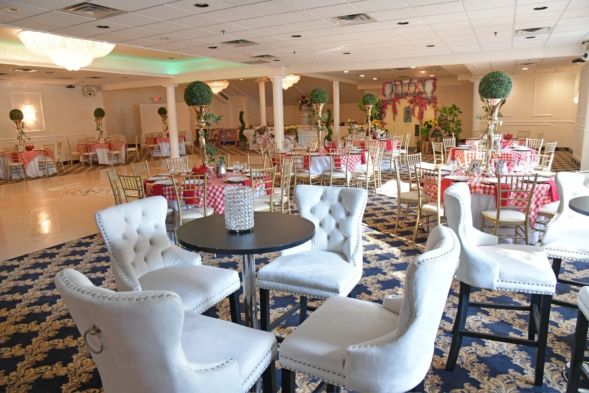 An elegantly decorated banquet hall with multiple round tables covered in red and white checkered tablecloths, gold chairs, and tall centerpiece vases with greenery. The front features white tufted chairs around small black tables, and the background