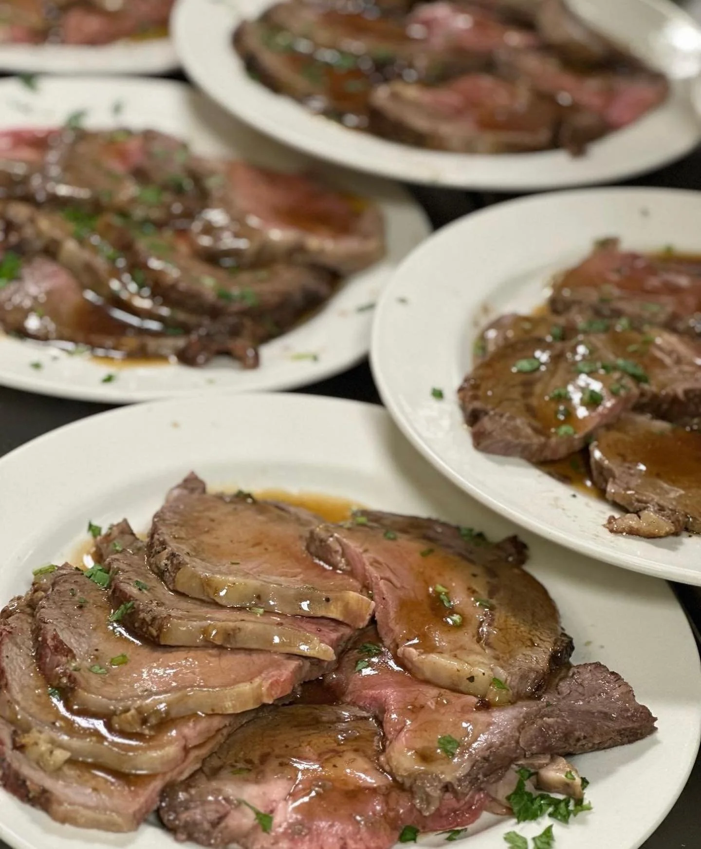 Several white plates filled with cooked beef steaks garnished with chopped parsley and topped with a brown sauce.