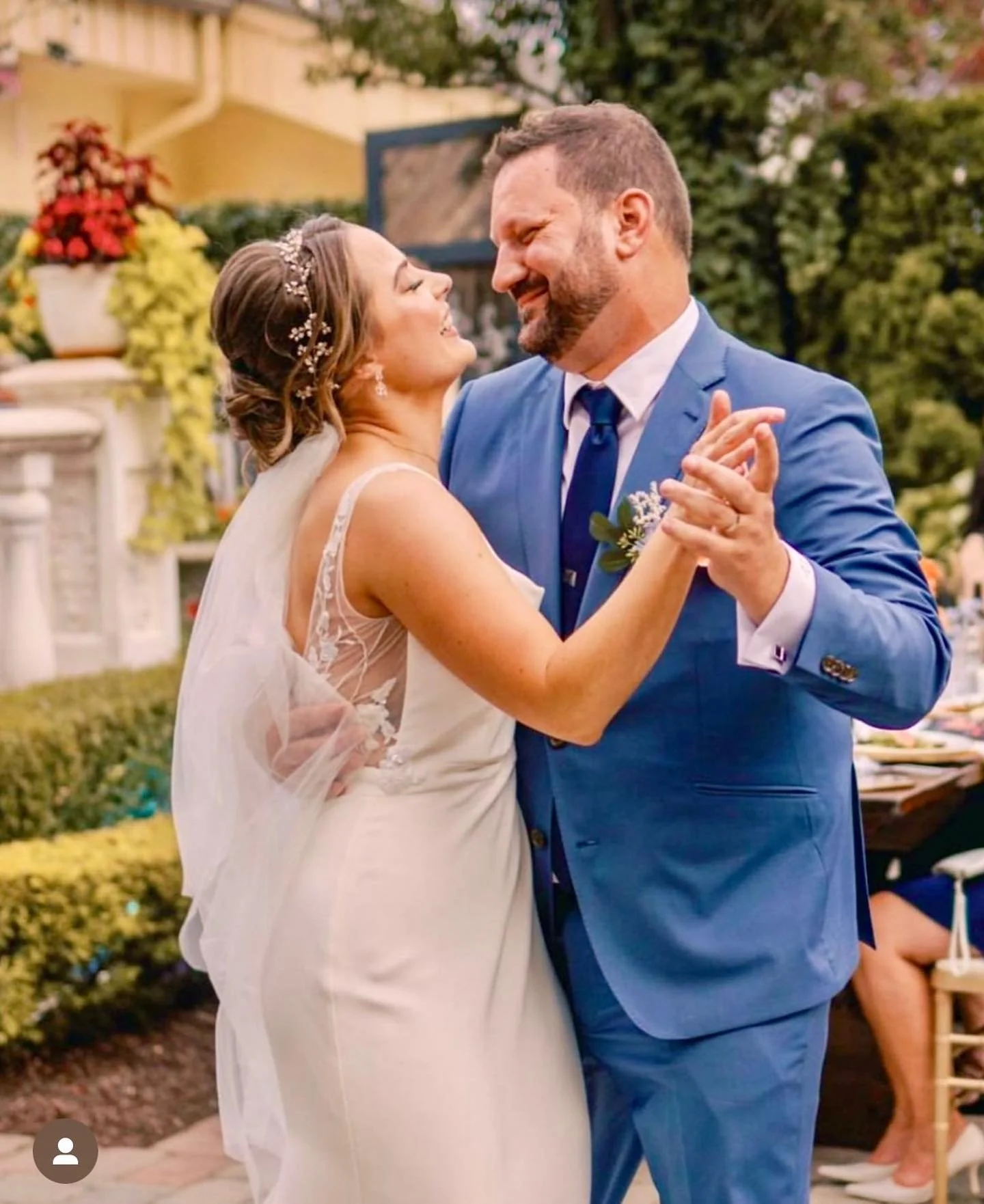 A bride and groom sharing a dance outdoors at their wedding, with the bride in a white gown and the groom in a blue suit, smiling at each other.