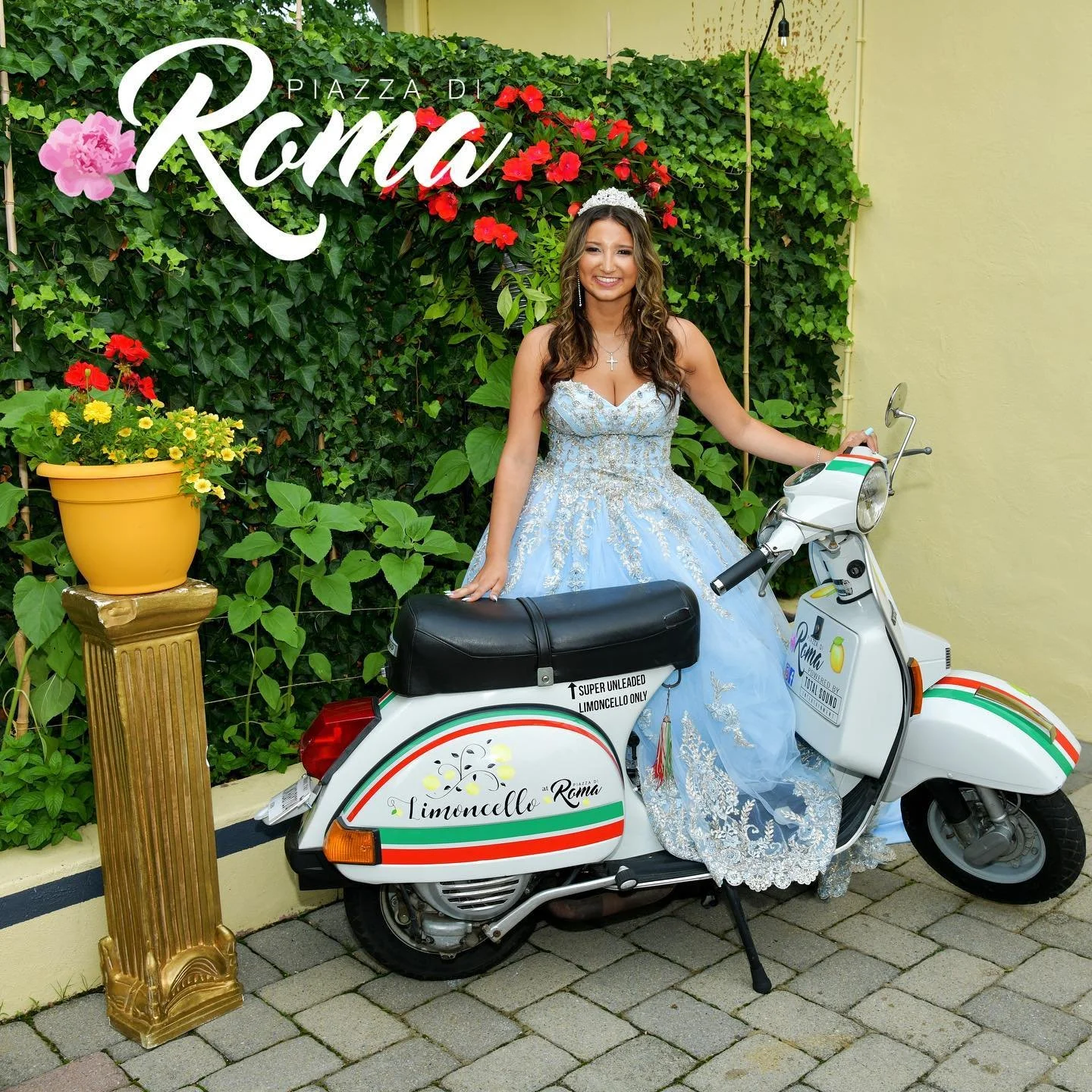 A young woman in a light blue wedding gown standing next to a white scooter with Italian flag colors, in front of a green ivy wall and a yellow wall. She is holding the scooter and smiling at the camera.