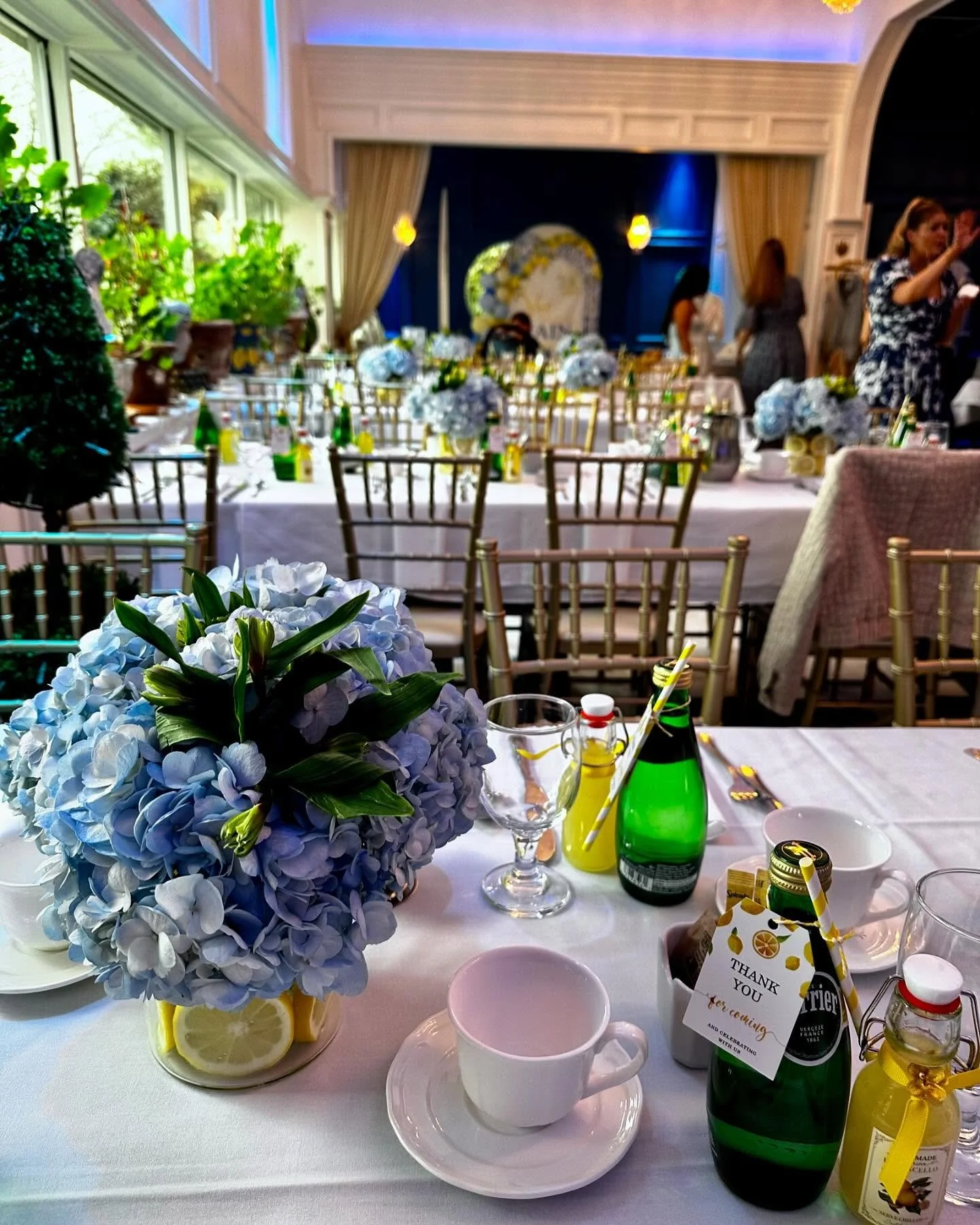 A wedding reception table set with blue hydrangea centerpieces, lemon slices, and assorted beverages, with chairs and other decorated tables in the background.
