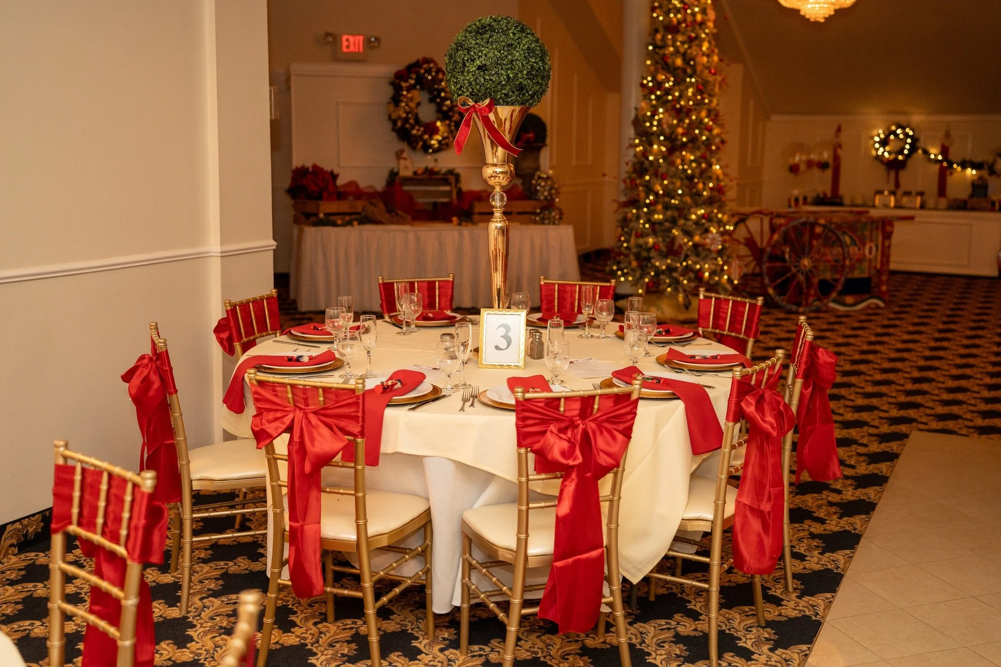 Festively decorated banquet table with gold chairs adorned with red bows, set with plates, glasses, and napkins, in a room with Christmas trees and wreaths.