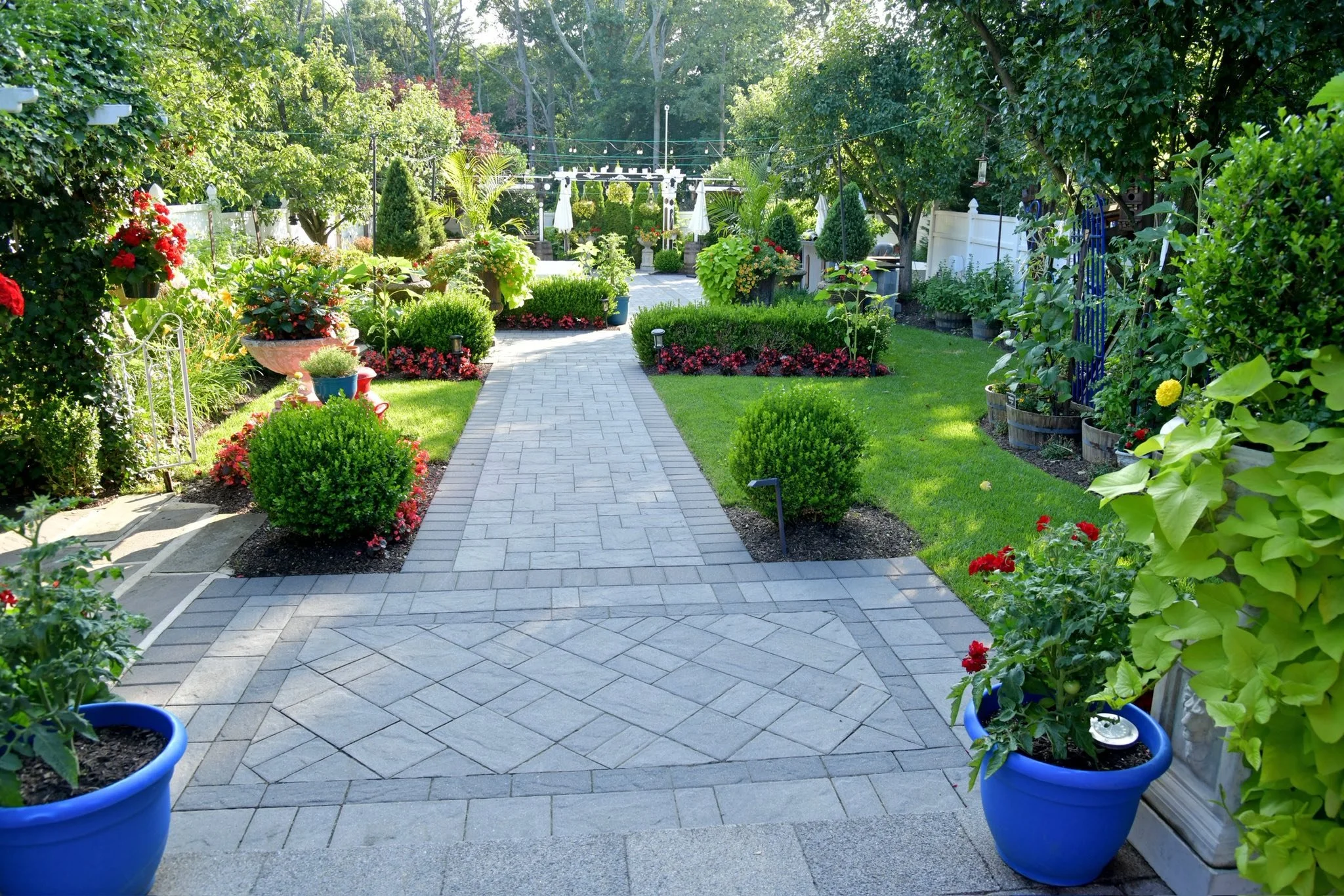 View of a well-maintained backyard garden with a paved pathway, colorful flowers, lush green plants, and trees.