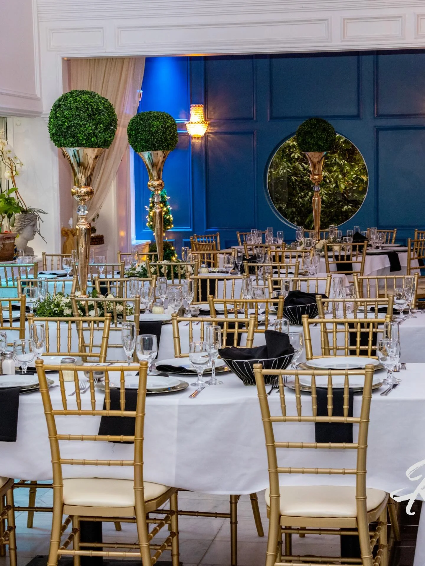 Elegant banquet hall with tables set for a formal event, featuring gold chairs, white tablecloths, black napkins, and tall decorative topiary plants, with a blue accent wall and a round mirror in the background.